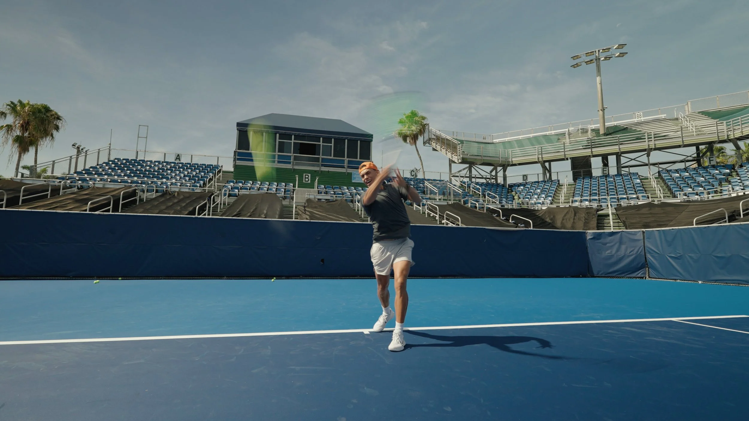 A person in athletic clothing and white shoes celebrating on a blue tennis court, with empty stadium seating and palm trees in the background during sunny weather.