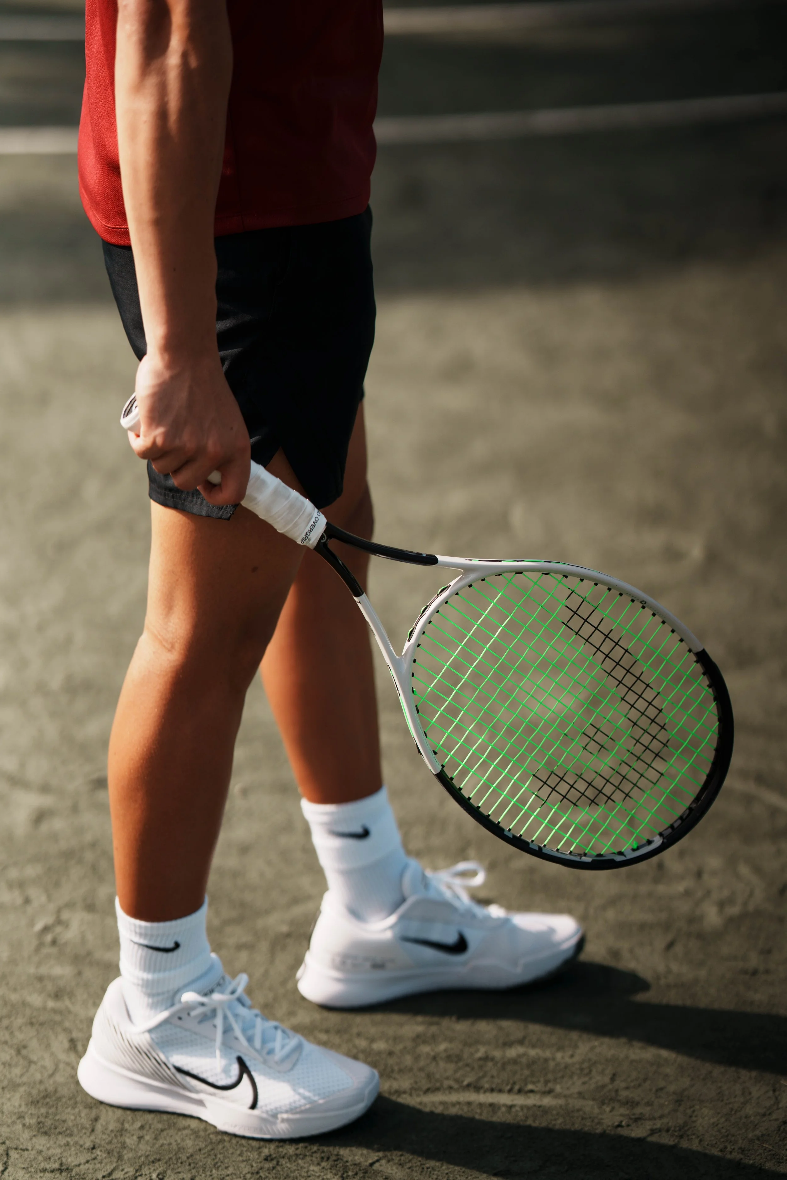 Close-up of a tennis player's lower body on a tennis court, holding a tennis racket, wearing white Nike sports shoes, white Nike socks, black shorts, and a red shirt.