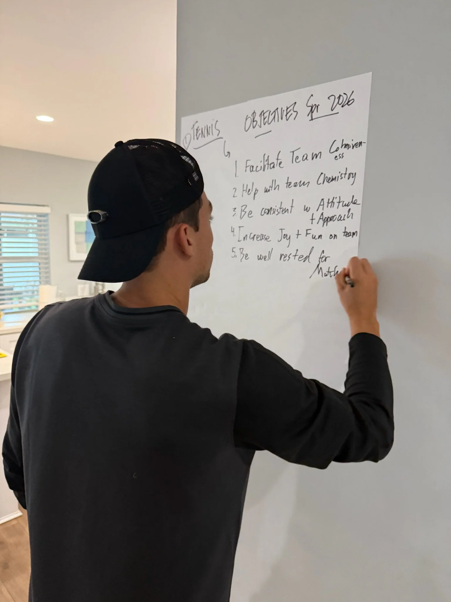 A person wearing a black baseball cap and black long-sleeve shirt writes on a white sheet of paper taped to a gray wall. The paper lists 'TENNIS OBJECTIVES for 2026' with five handwritten goals.