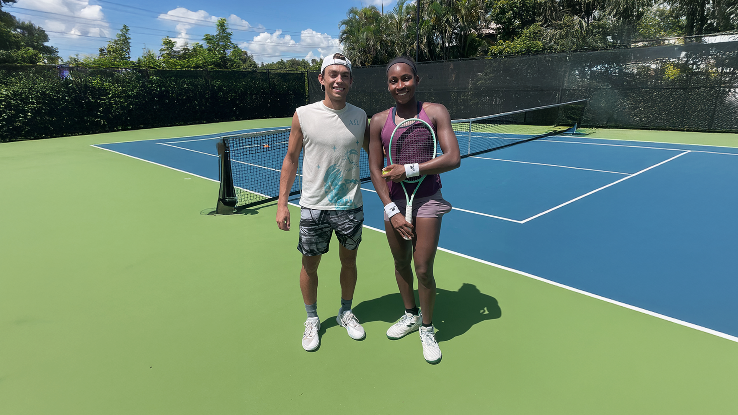 Two people standing on a tennis court, smiling. One is a man wearing a white sleeveless shirt, patterned shorts, and a white cap. The other is a woman wearing a purple tank top, shorts, and holding a tennis racket. There is a tennis net behind them, with greenery and a partly cloudy sky in the background.