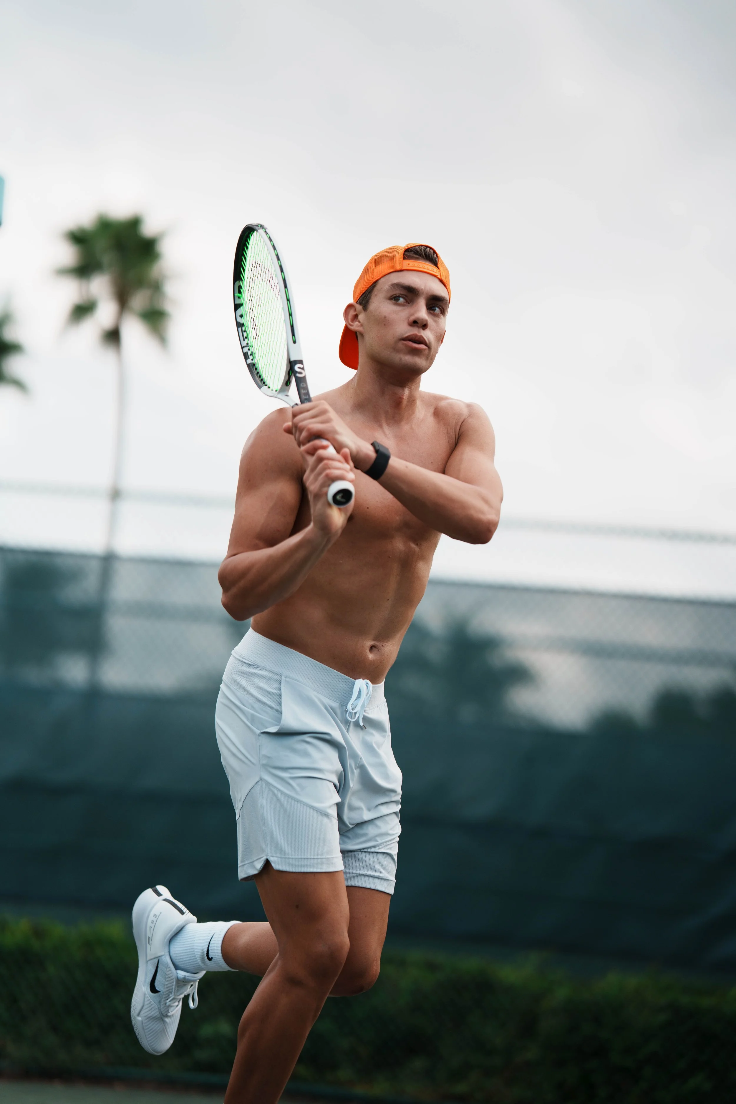 A young man with a fit physique and light skin, wearing a backward orange cap, white shorts, and white Nike sneakers, is playing tennis on an outdoor court during daytime. He is holding a tennis racket in both hands and appears focused. The background shows a chain-link fence and trees under an overcast sky.