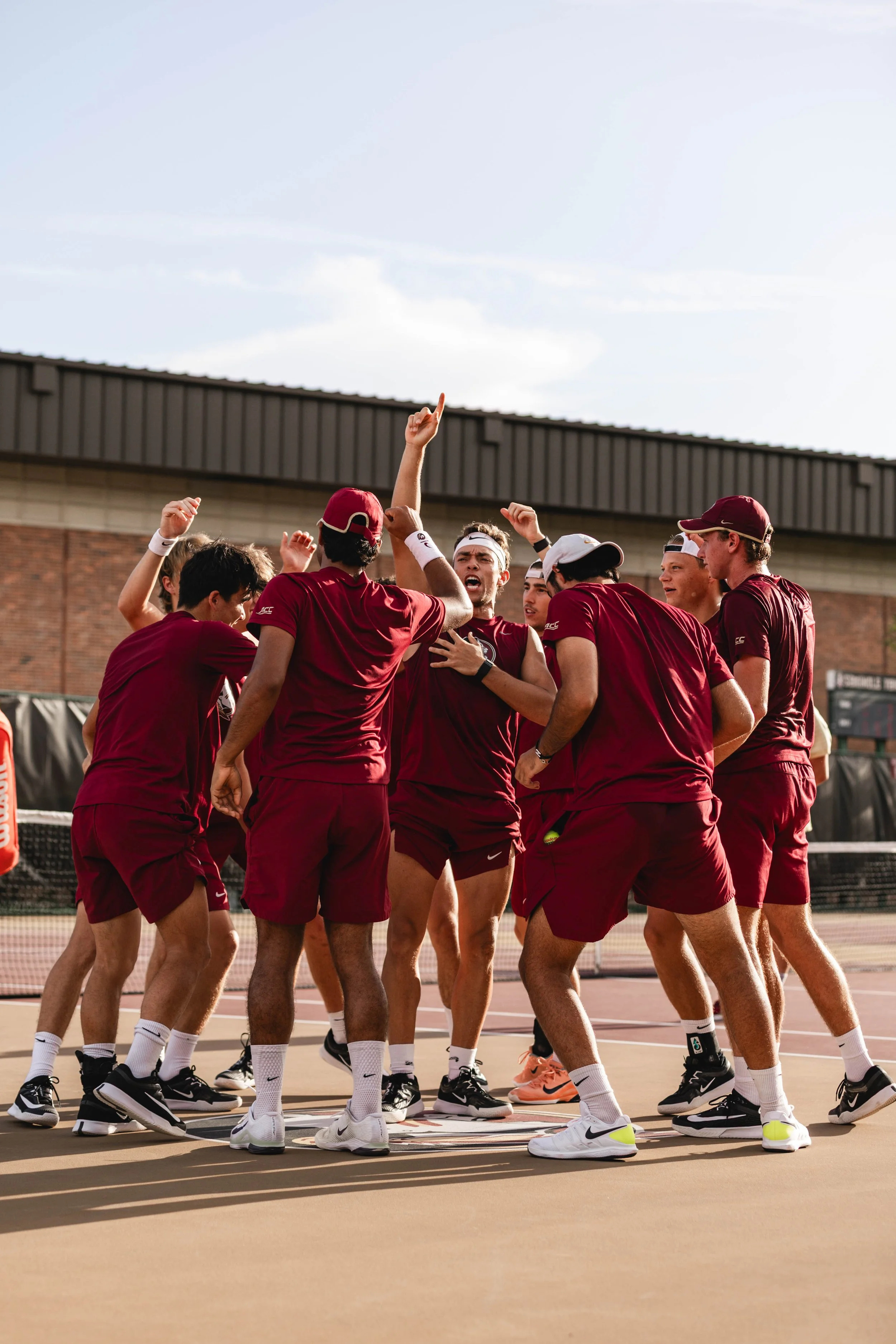 College tennis team in matching maroon uniforms huddles in a circle on an outdoor tennis court, with a brick building and blue sky in the background.