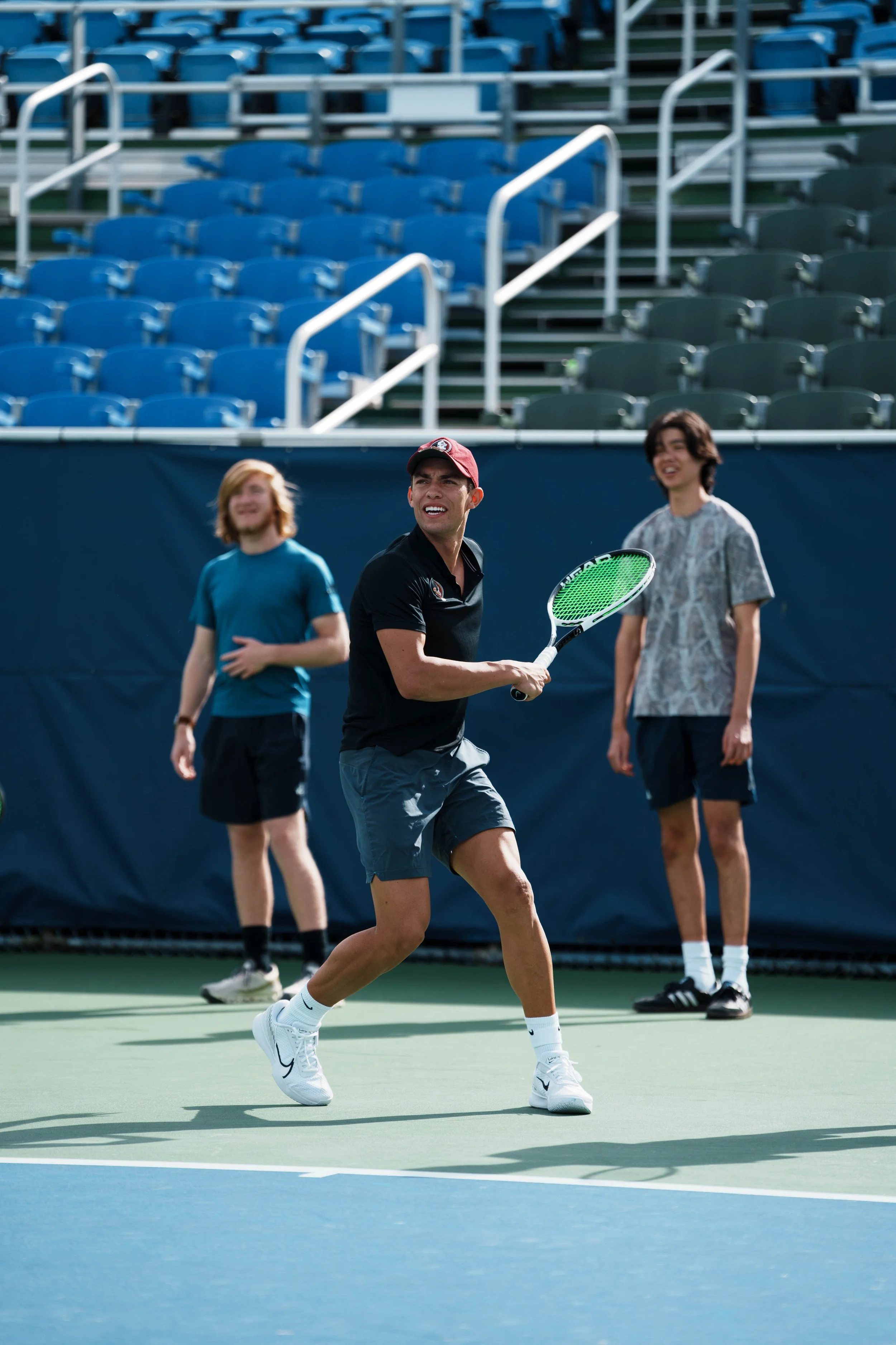 A man playing tennis on an outdoor court with two other men in the background, one wearing a teal shirt and the other in a patterned shirt, all standing in front of empty blue and green stadium seating.