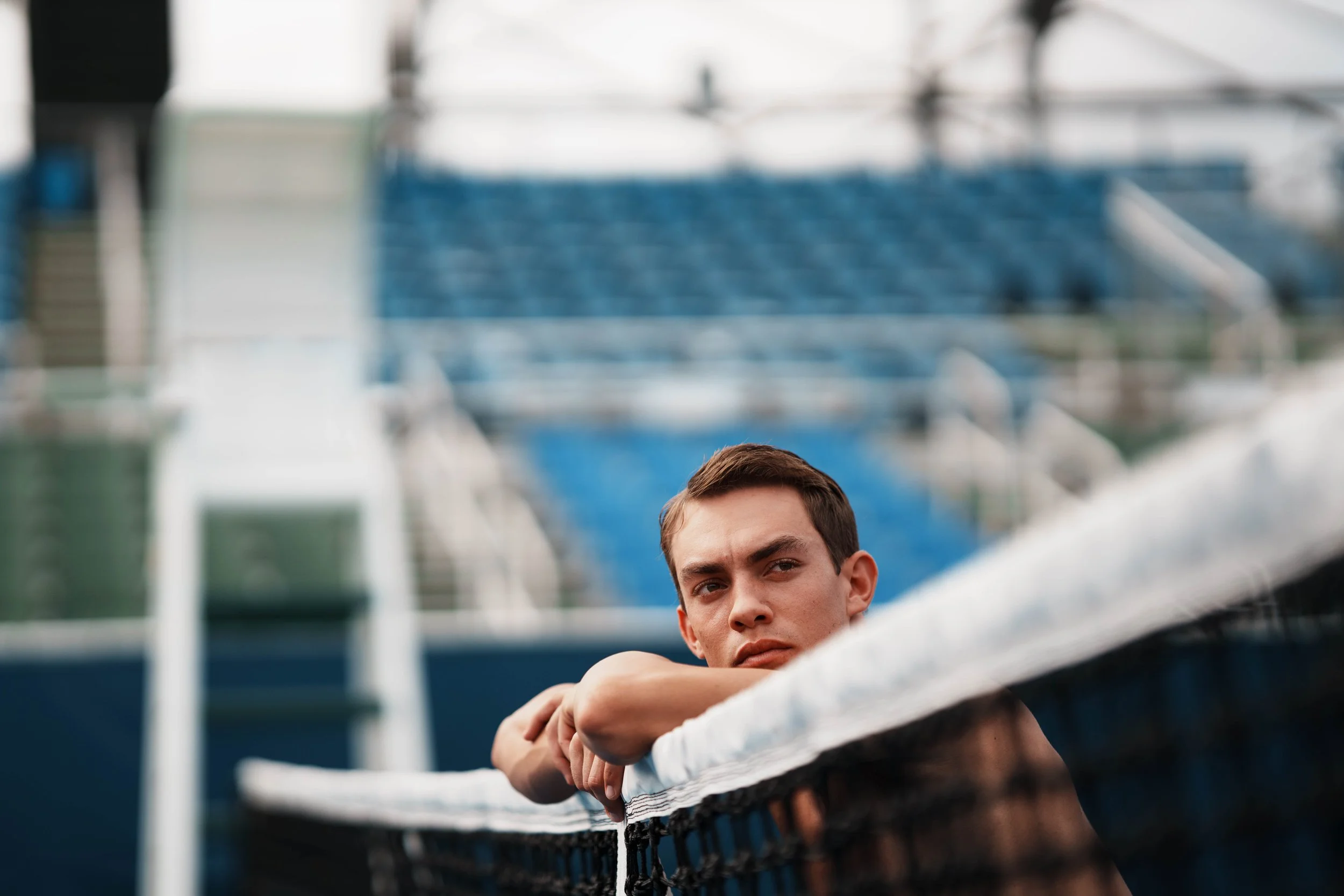 Young man with short hair and a serious expression leaning on a white railing at a sports stadium, with blue seats and part of the stadium structure visible in the background.