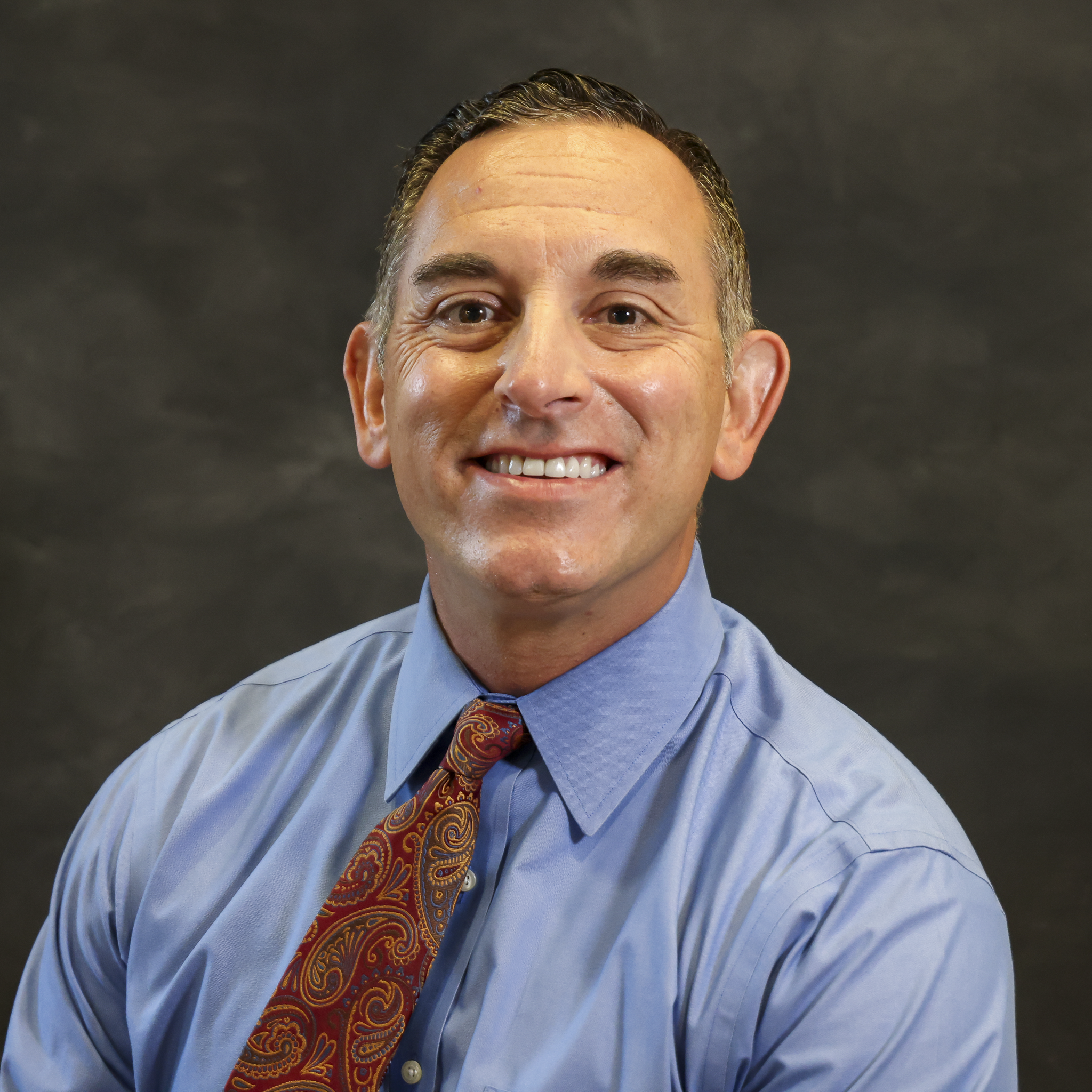 Close-up of a man wearing a blue dress shirt and a colorful paisley tie, smiling with his mouth slightly open, with a dark background.