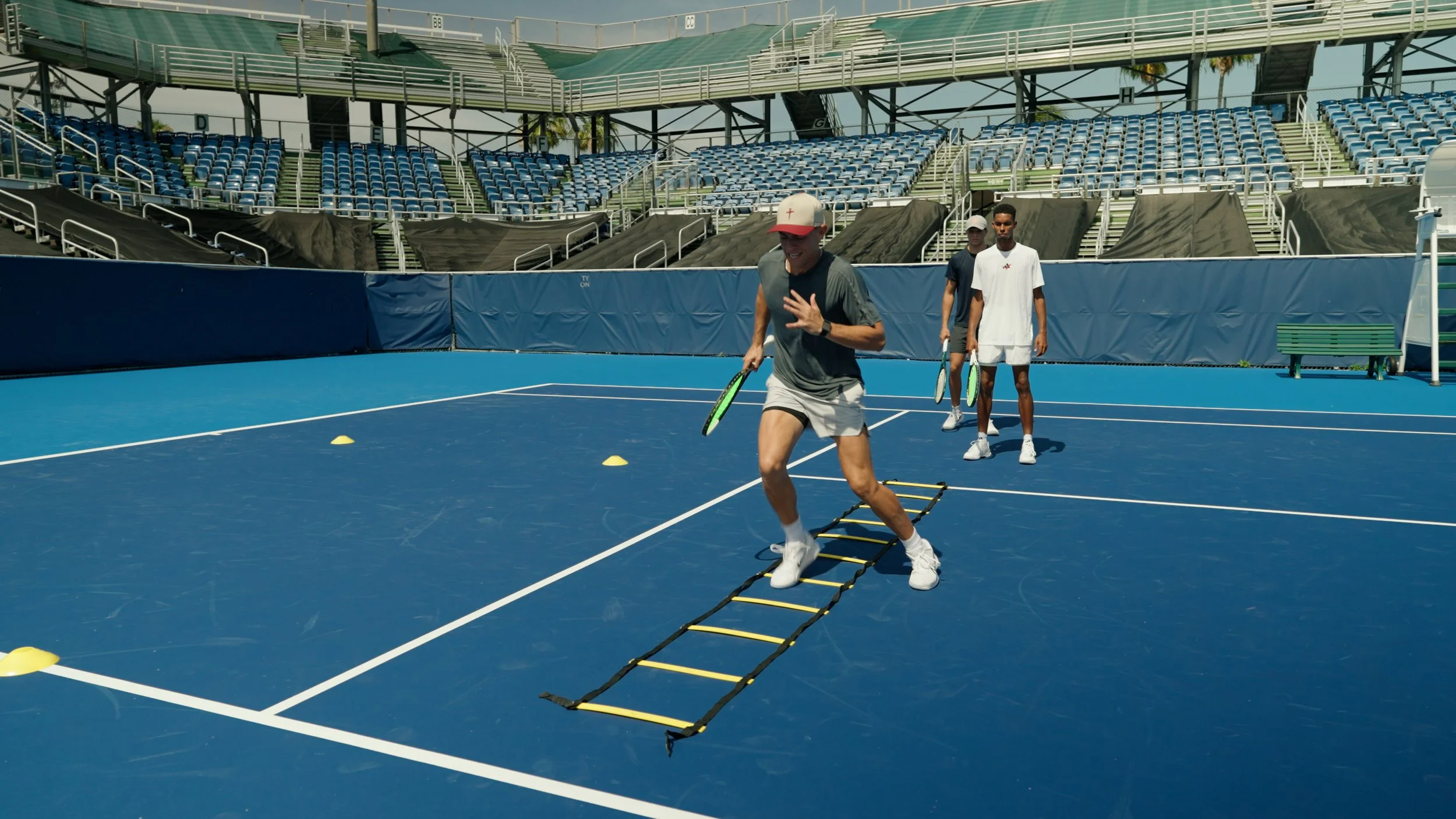 Three men practicing tennis drills on an outdoor blue tennis court, with stands and empty seats in the background.
