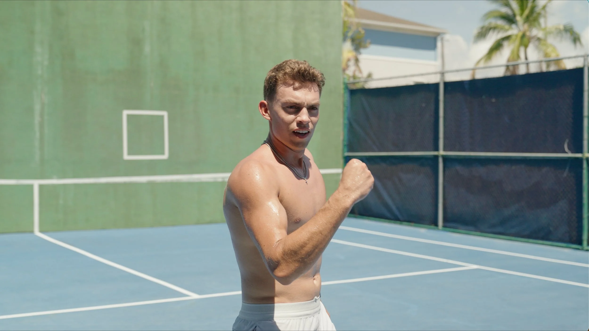 A shirtless young man with short brown hair, clenched fists, and a determined expression on his face, standing on an outdoor tennis court with palm trees and a blue sky in the background.