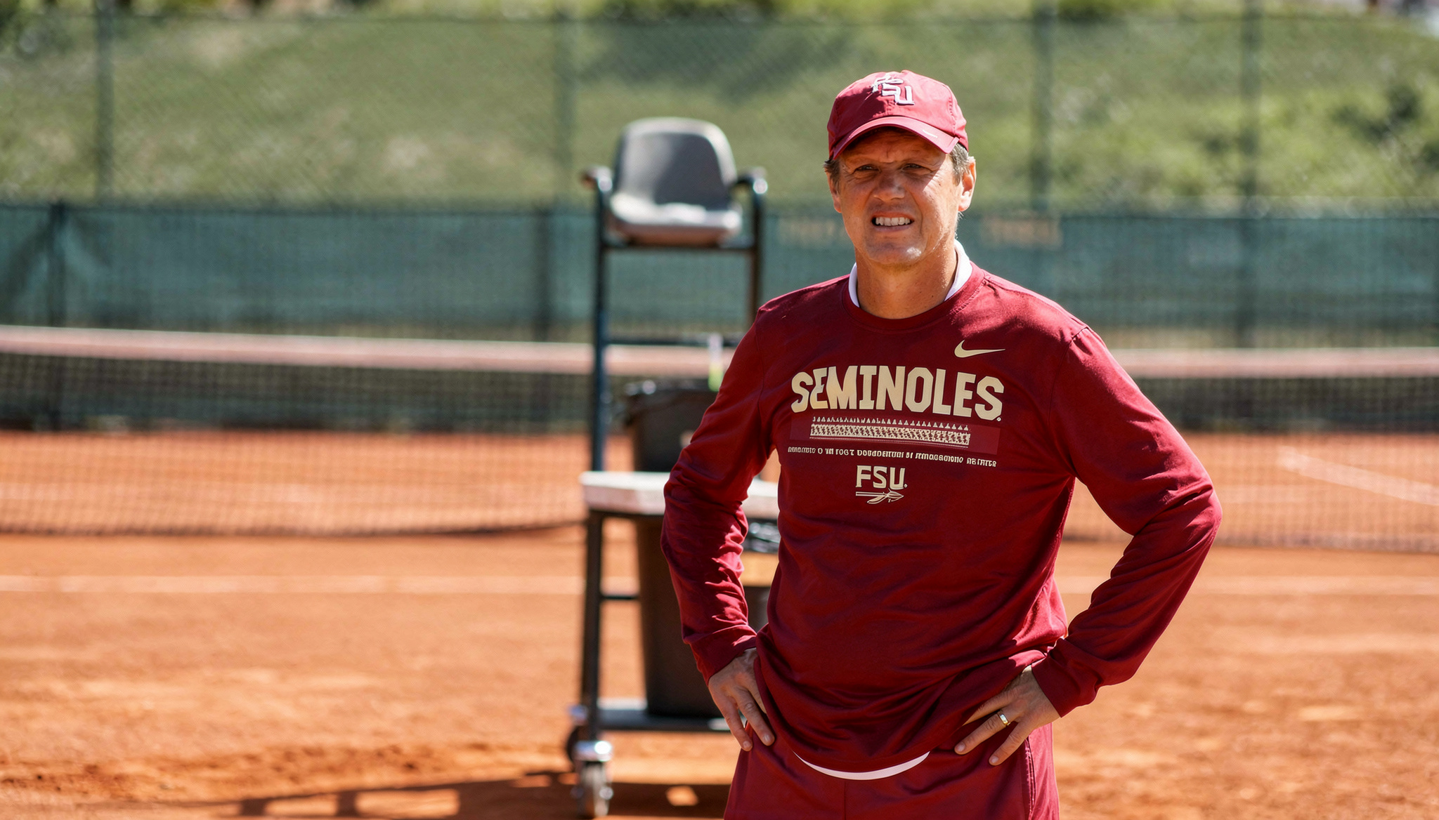 A man standing on a clay tennis court wearing a red cap and red shirt with 'Seminoles' printed on it. He appears to be focused or concerned, with outdoor tennis court equipment visible in the background.