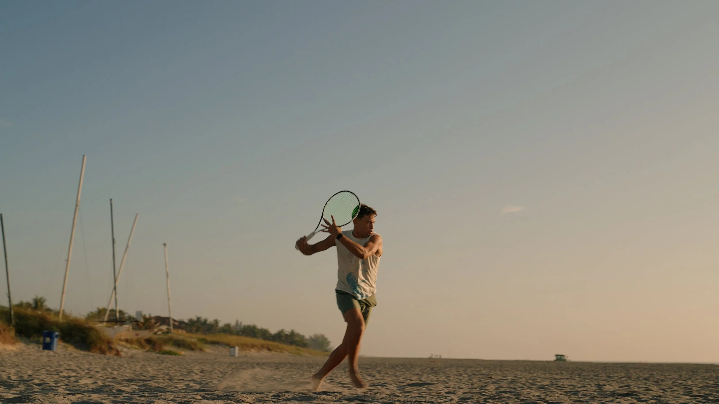 A young man playing tennis on a beach during sunset.