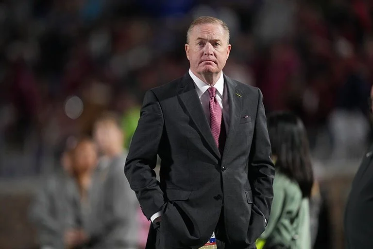 A man in a black suit and red tie stands with his hands in his pockets at a public event, with a blurred crowd in the background.