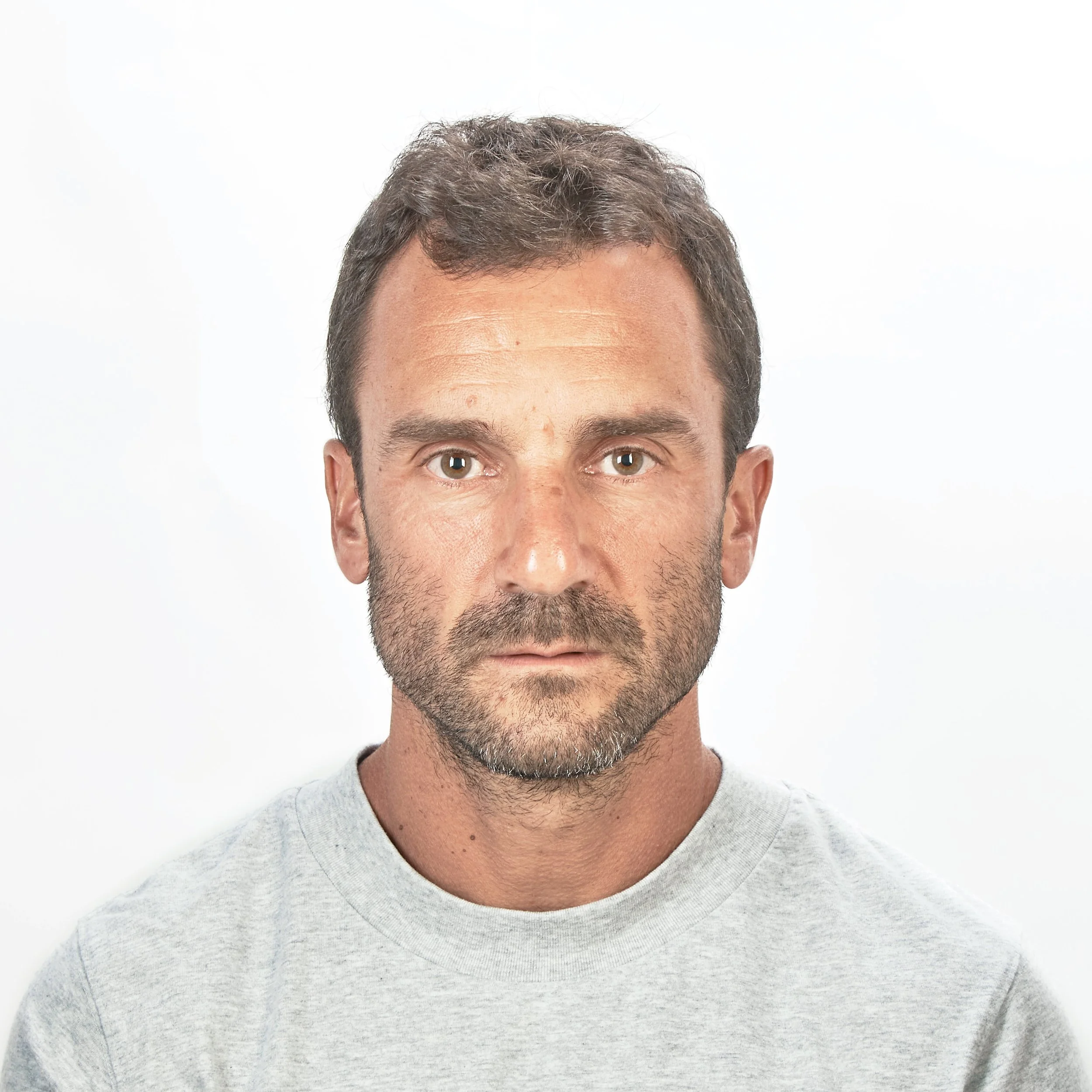 Close-up of a man's face with brown hair and brown eyes, wearing a light gray sweater, against a plain white background.