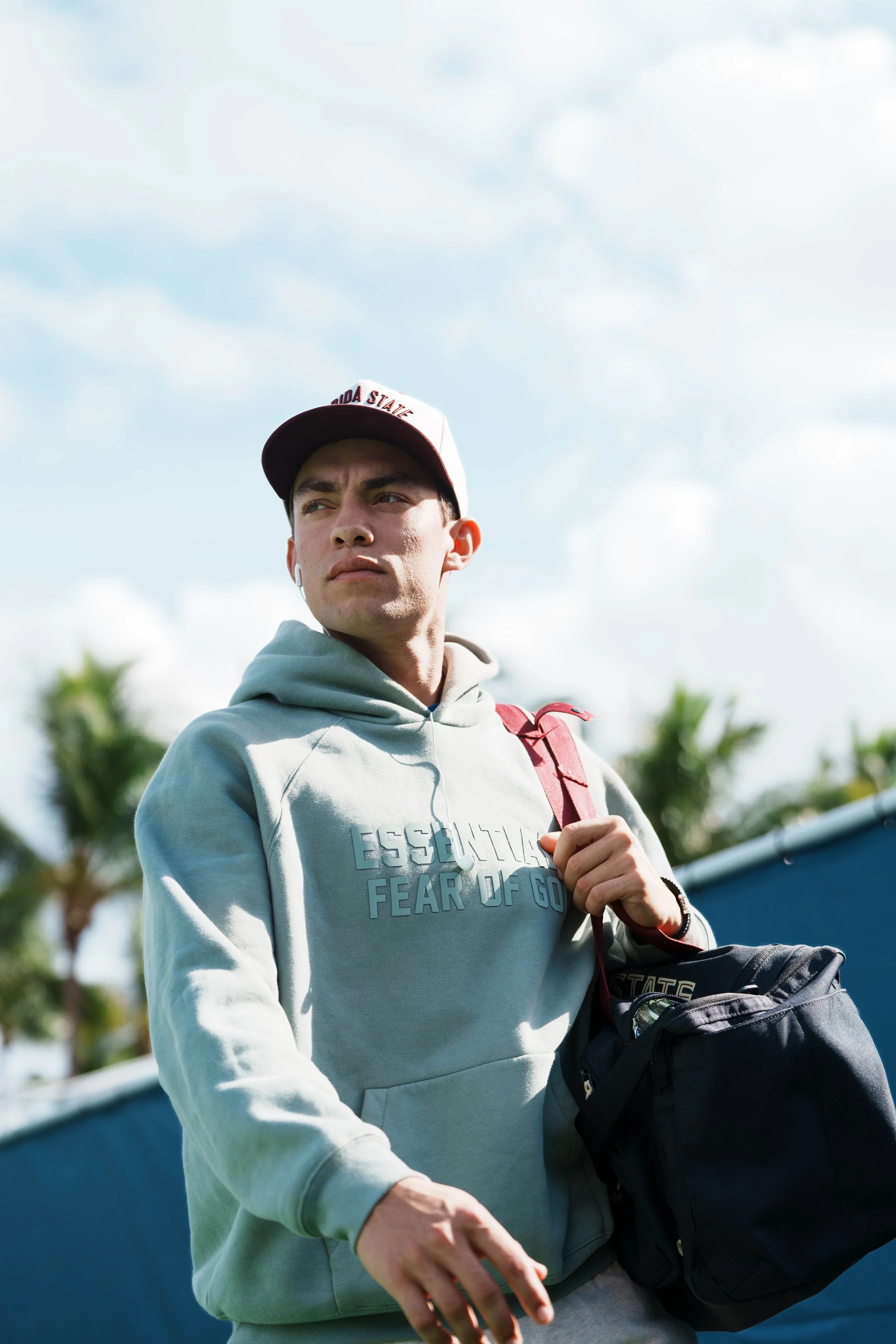 A young man wearing a white and red Ohio State cap, a gray hoodie with text, and carrying a black backpack, outdoors on a partly cloudy day with palm trees in the background.