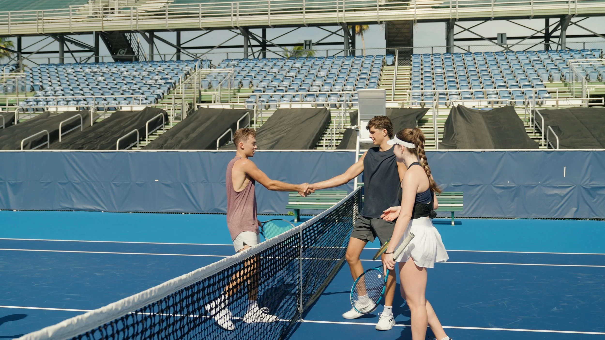 Three tennis players shaking hands at the end of a match on an outdoor tennis court with empty bleachers in the background.