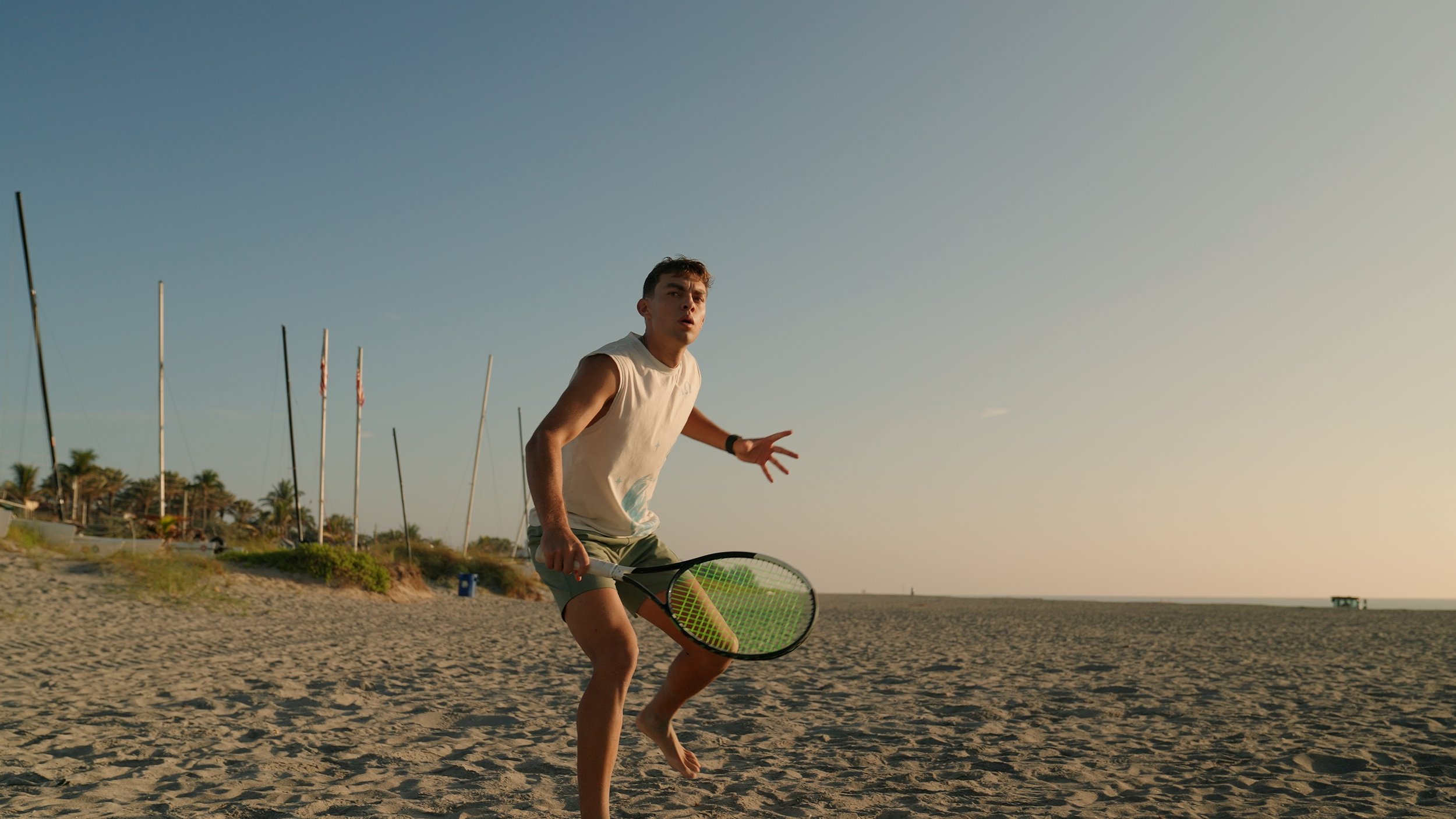 A young man playing tennis on the beach during sunset, holding a tennis racket and wearing a sleeveless white shirt and shorts.