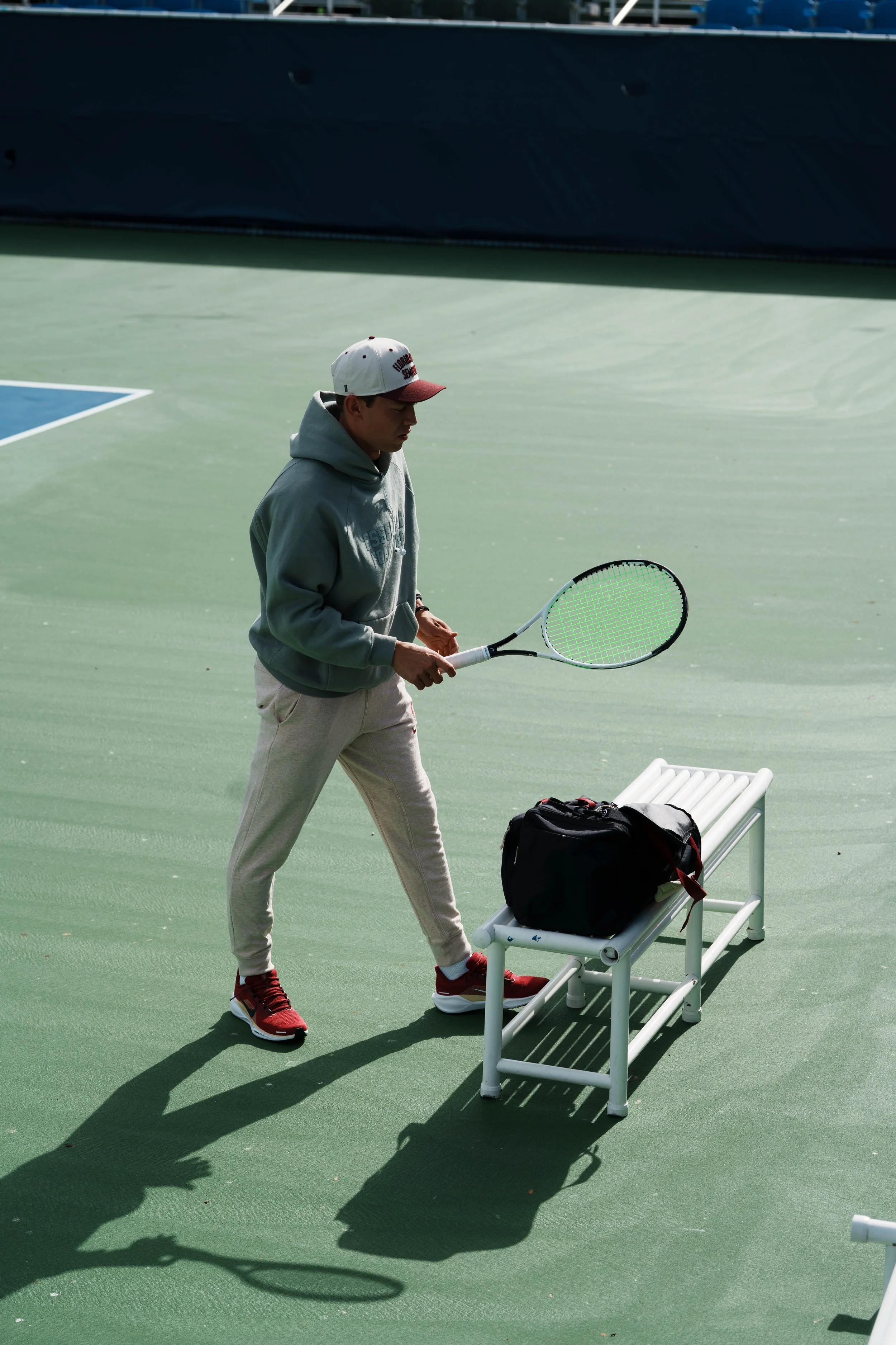Young man in gray hoodie and red sneakers holding a tennis racket, standing near a bench with a black bag on it, on an outdoor tennis court.