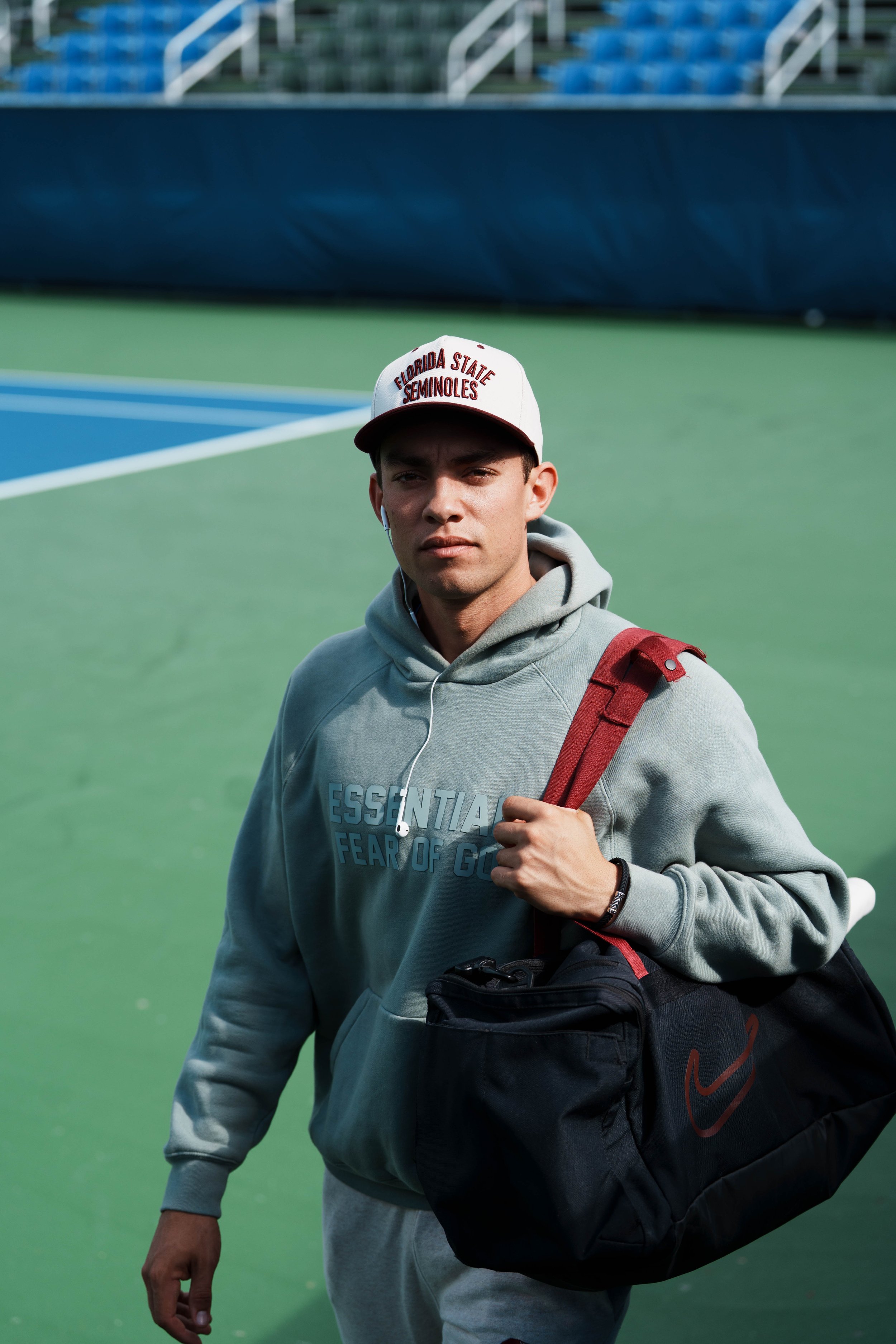 A young man walking on a tennis court, carrying a black and red backpack and wearing a white cap with red lettering that reads 'Florida State Seminoles'. He has earphones in and is dressed in a light grey hoodie with blue text and grey sweatpants.