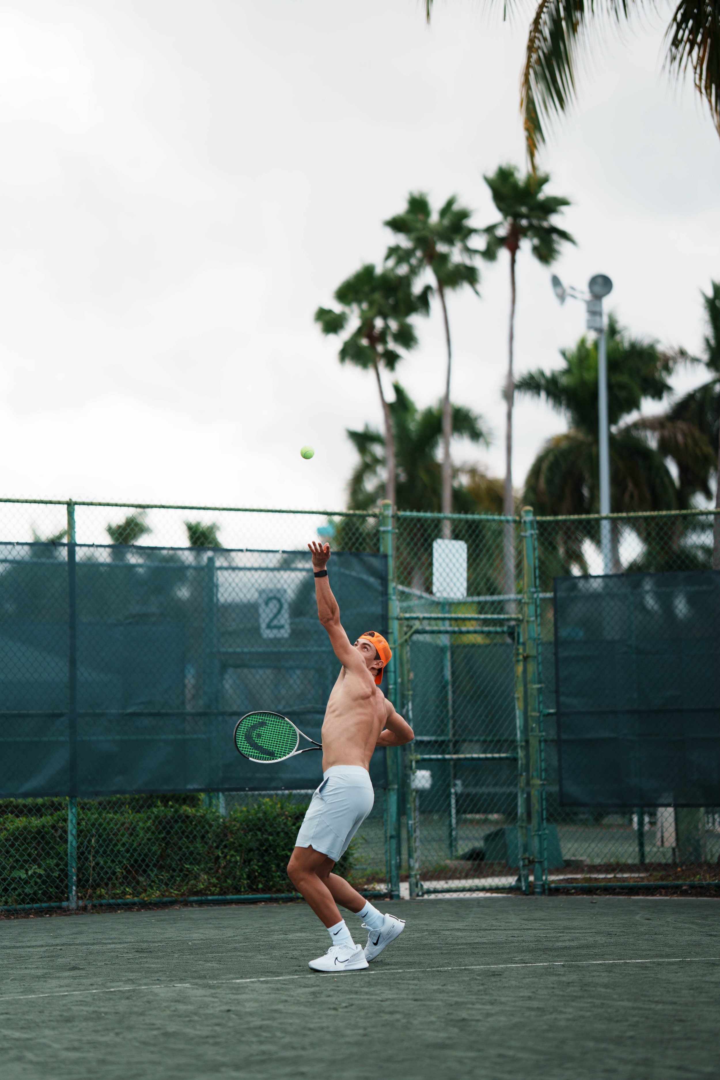 A shirtless tennis player wearing white shorts, white athletic shoes, and an orange headband is on a tennis court, swinging a green racket towards a tennis ball in mid-air. There are palm trees, a fence, and a cloudy sky in the background.