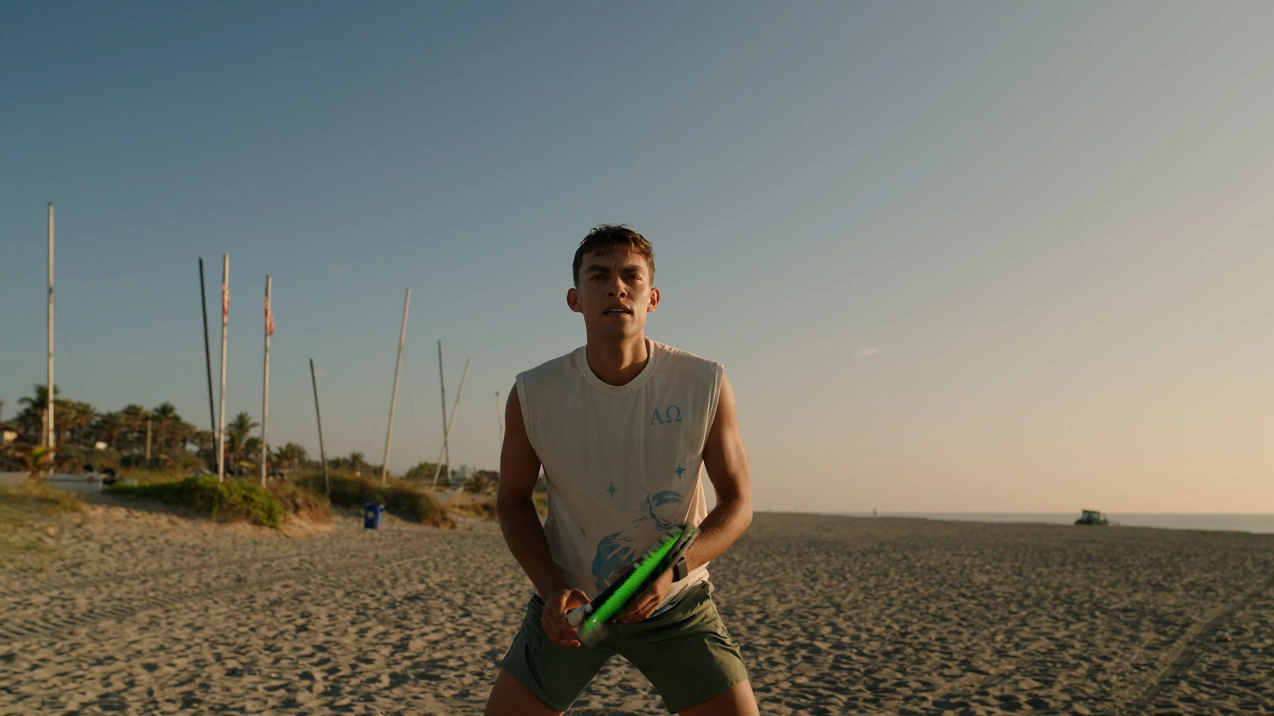 A young man on a beach holding a tennis racket, wearing a sleeveless shirt and shorts, with sailboats and houses in the background during sunset.