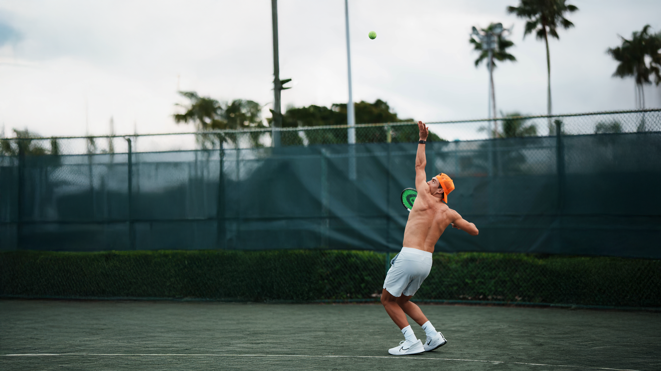 A shirtless young man wearing white shorts, white sneakers, and an orange cap is playing tennis on a court with a green surface, preparing to hit a tennis ball volleyed in the air.