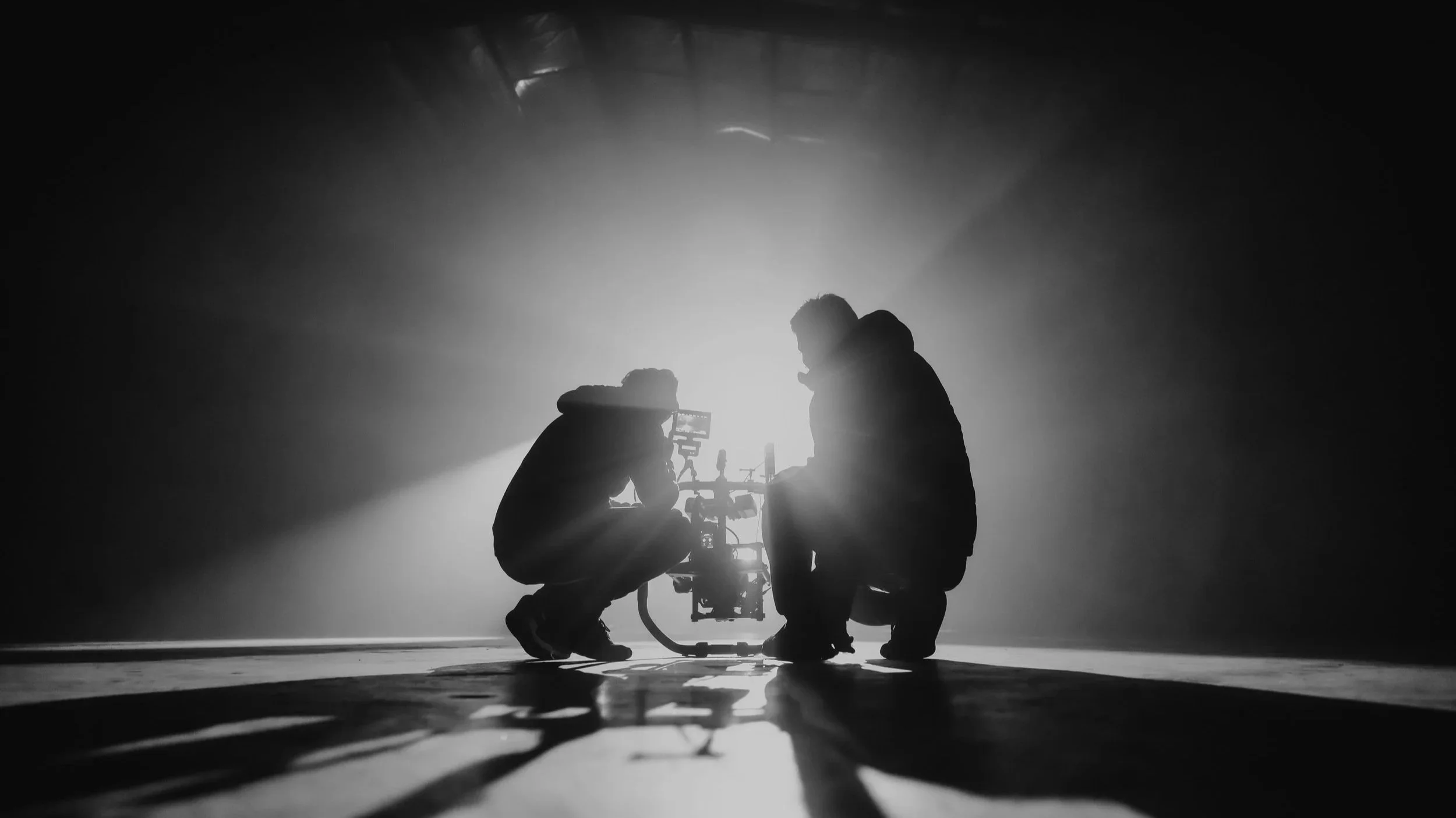 Two people in dark clothing are kneeling and working on a robot on the floor, with bright light shining behind them creating a silhouette and casting long shadows.