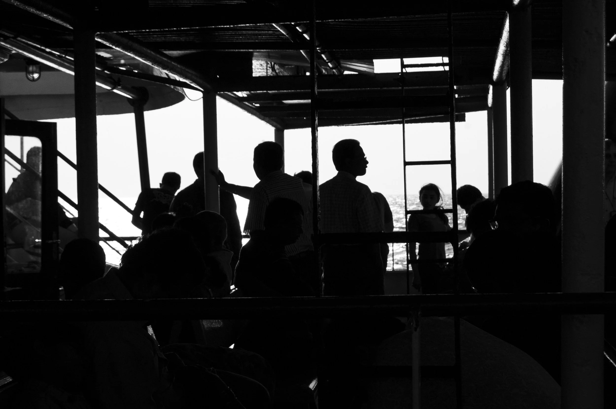 Silhouettes of people standing and sitting on a boat's upper deck, with the ocean in the background and sunlight shining through the open windows.