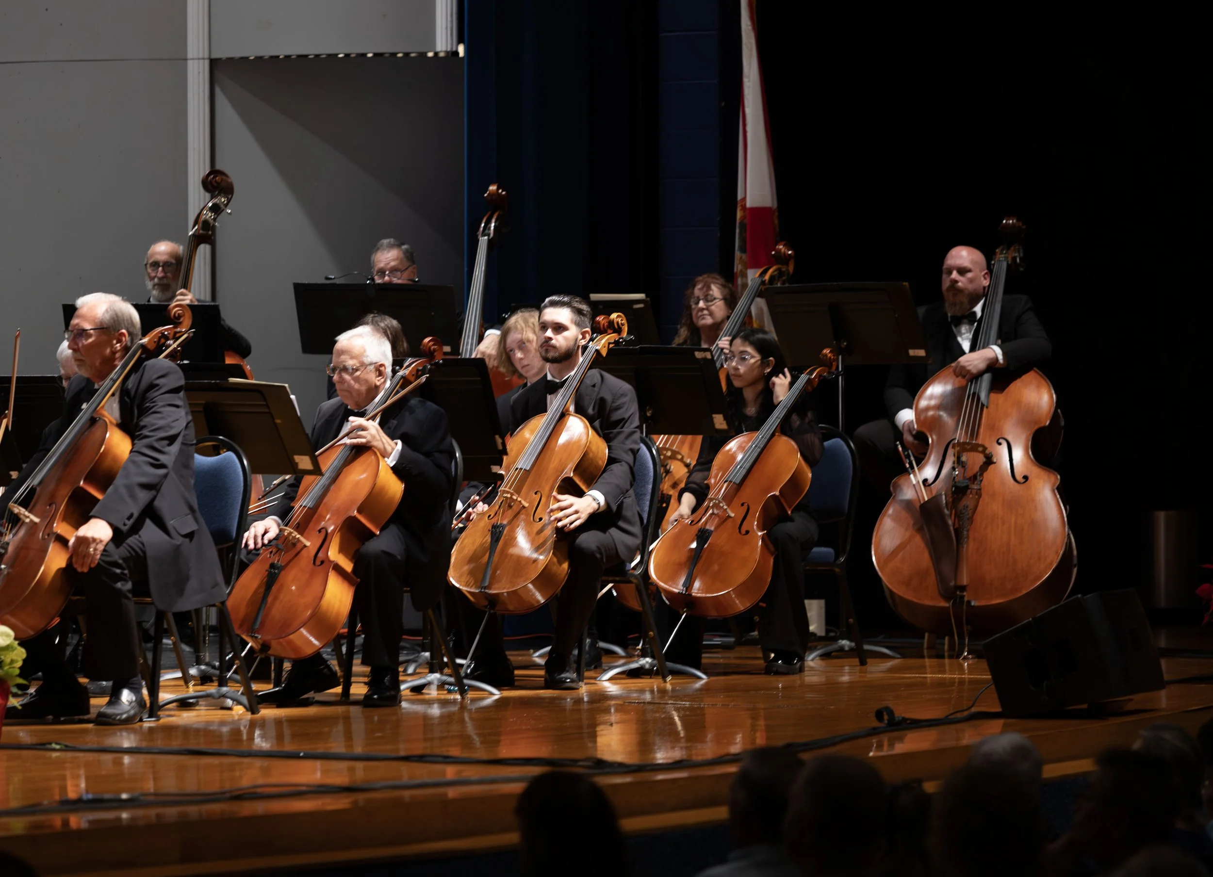 Orchestra performing on stage with string instruments including cellos, with a dark background and American flag in the background.