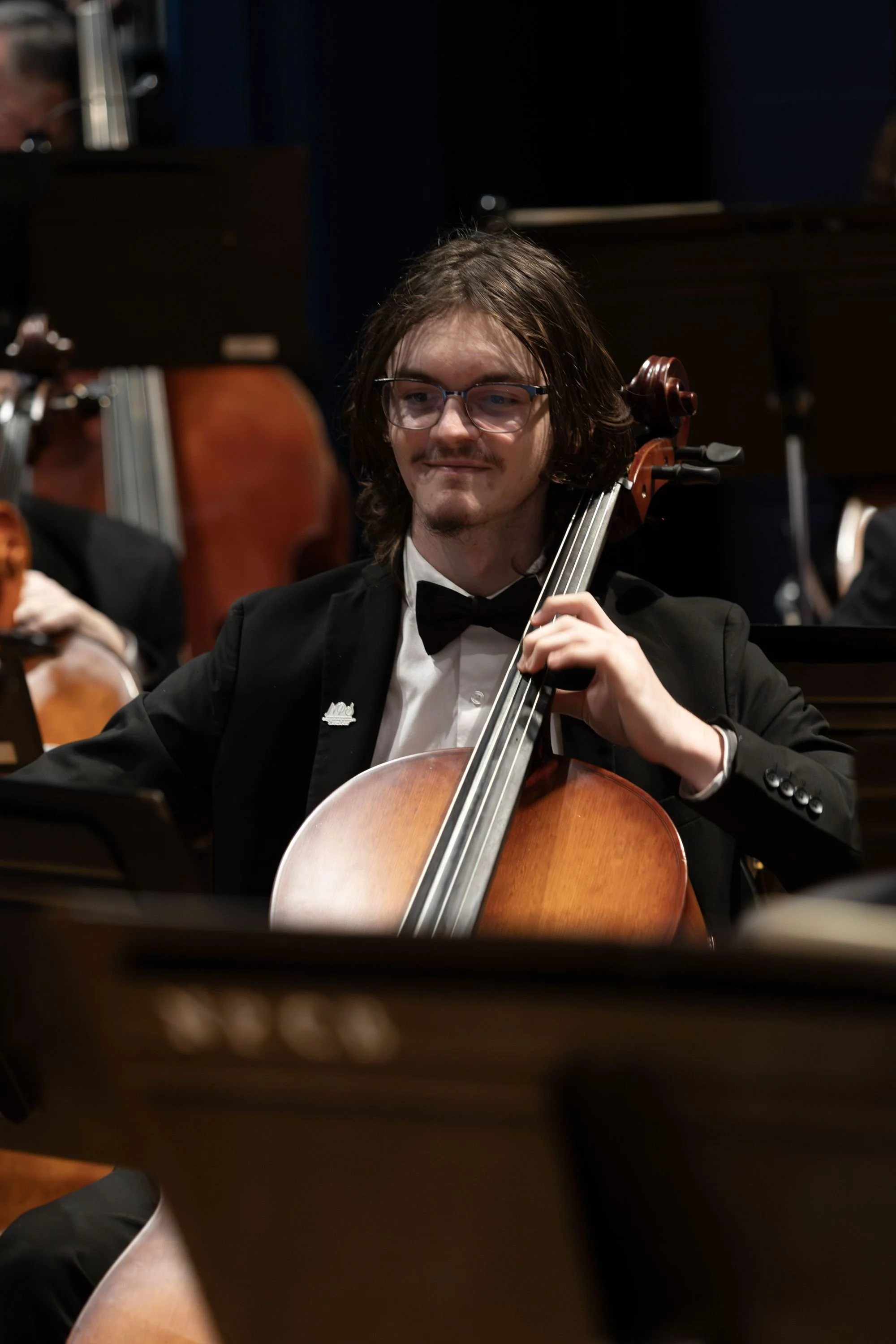 Young man with glasses and long hair playing the cello during an orchestra performance, dressed in a tuxedo.