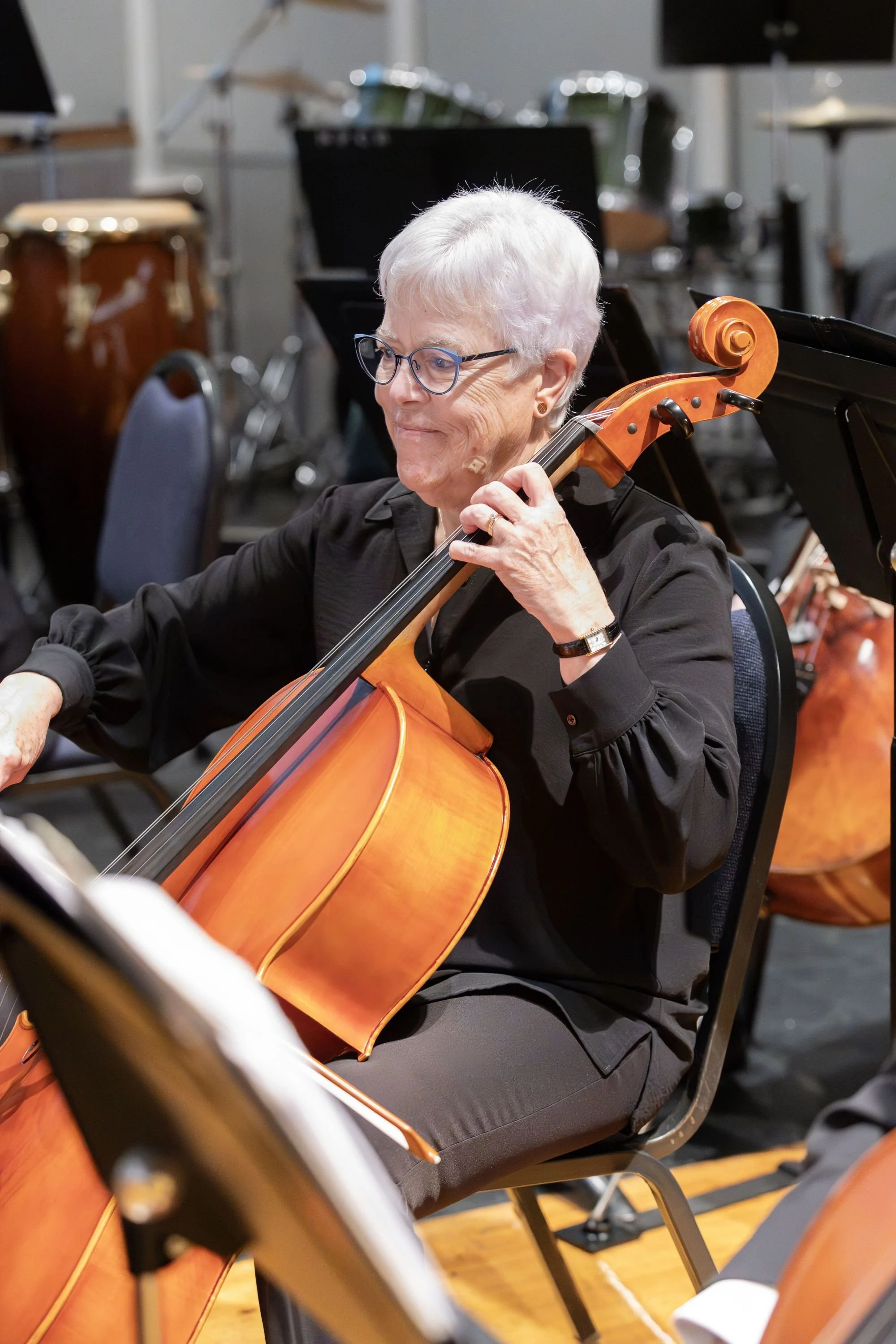 An elderly woman with short white hair, glasses, and wearing a black shirt, playing a wooden cello during a musical performance with percussion instruments in the background.