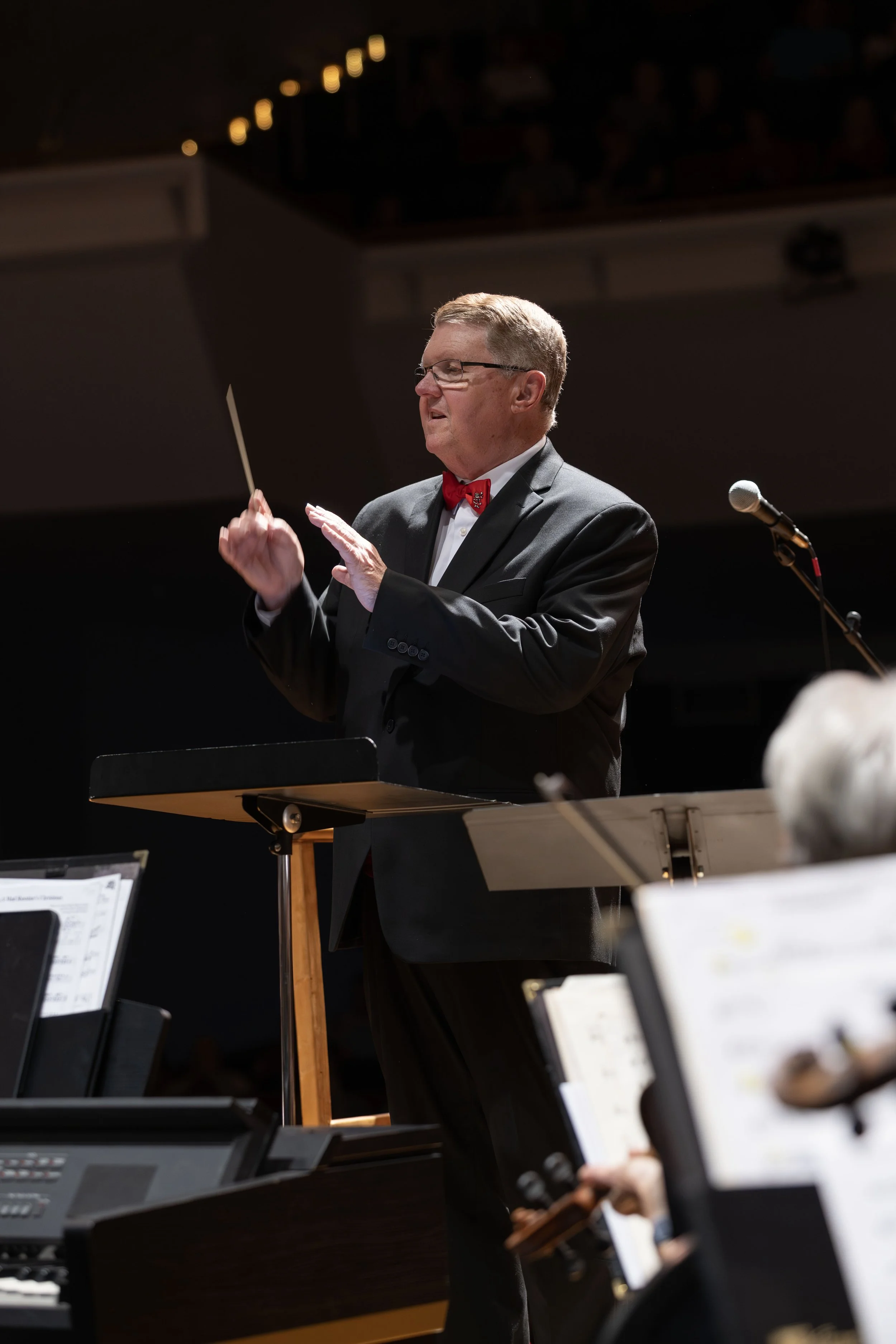 A man in a black tuxedo with a red bow tie conducting an orchestra in a concert hall.