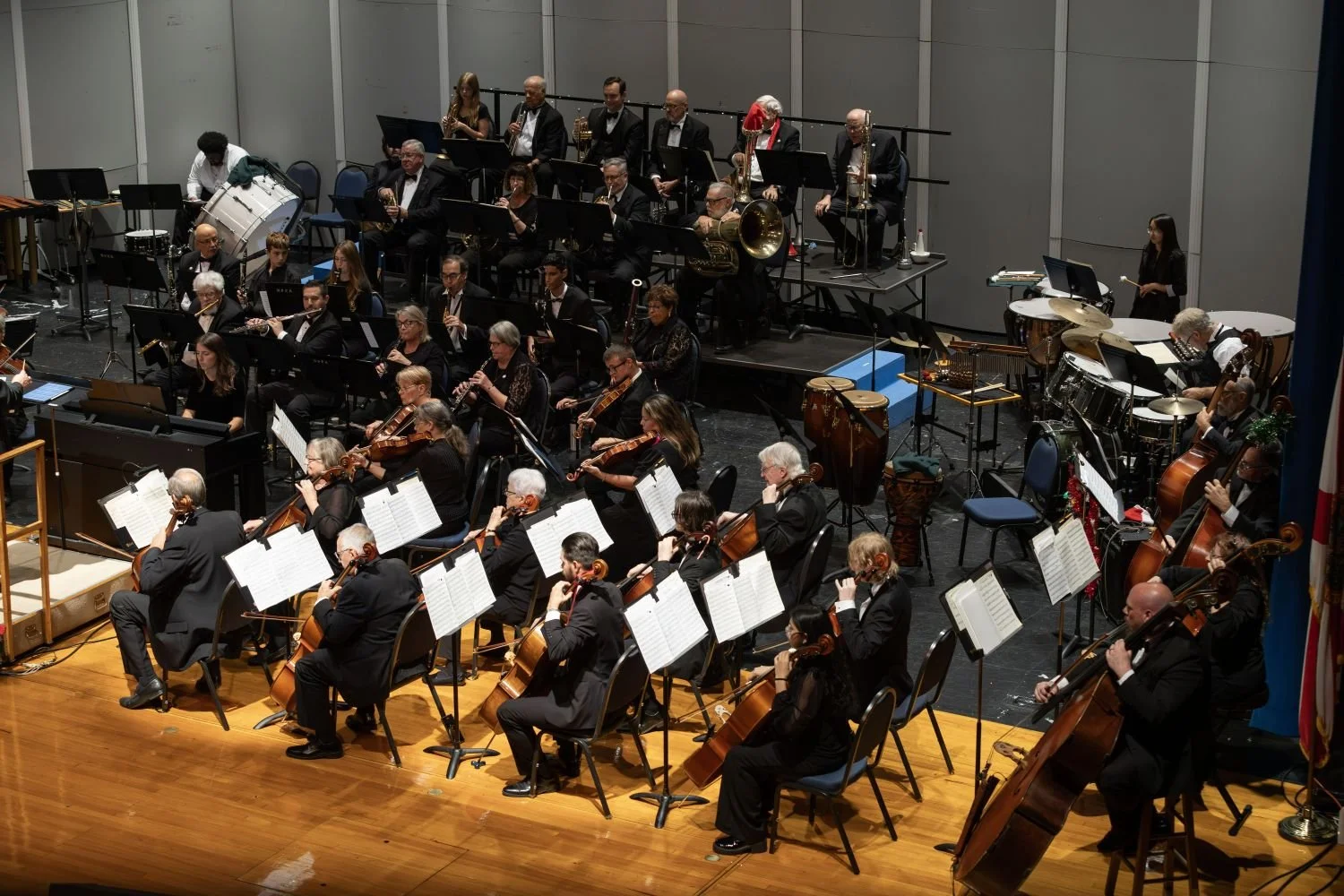 An orchestra performing on stage with musicians playing various instruments including violins, cellos, drums, and wind instruments, in a concert hall.