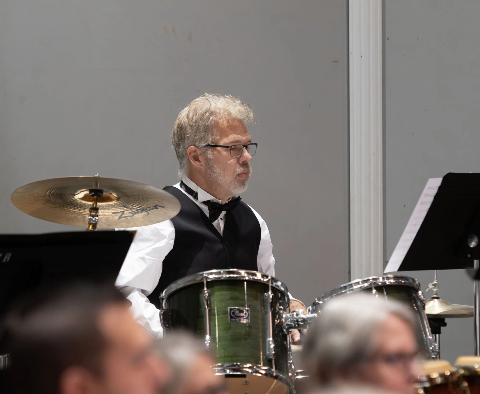 An older man with gray hair and glasses playing drums in an orchestra, wearing a black vest, white shirt, and black bow tie, with others in the background.