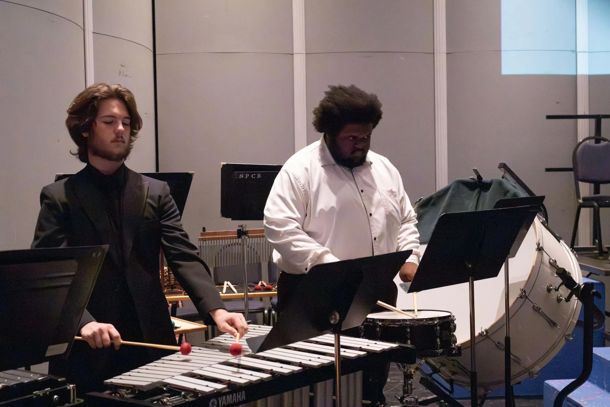 Two musicians playing percussion instruments, one striking a xylophone and the other playing a timpani, during a performance in a concert hall.