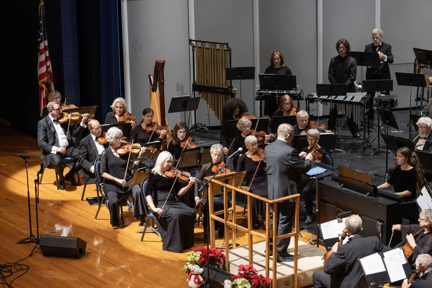 An orchestra performing on stage with a conductor, with musicians playing violins, cellos, and percussion instruments, all dressed in formal black attire, during a concert or event.
