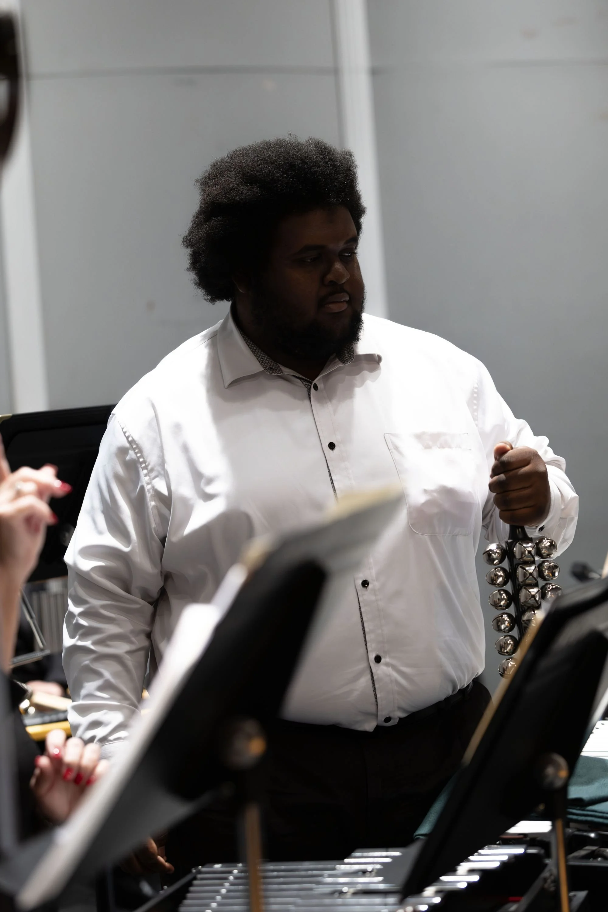 A man with an afro hairstyle and beard wearing a white dress shirt holding a percussion instrument with bells in a music rehearsal room.