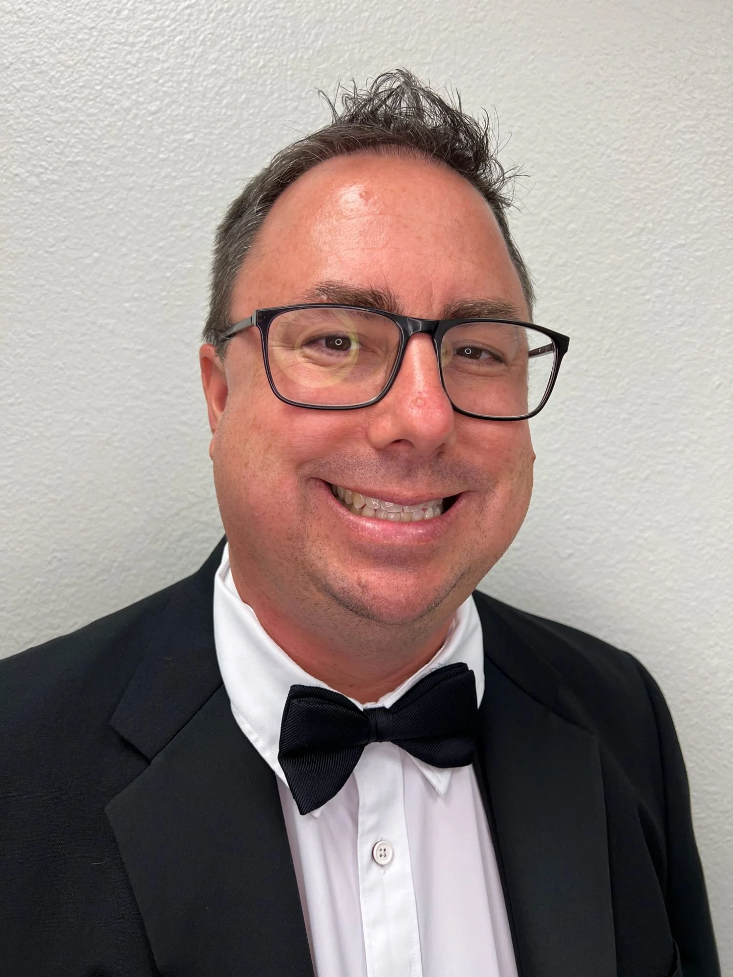 A man wearing glasses, a tuxedo with a bow tie, smiling, against a plain wall.