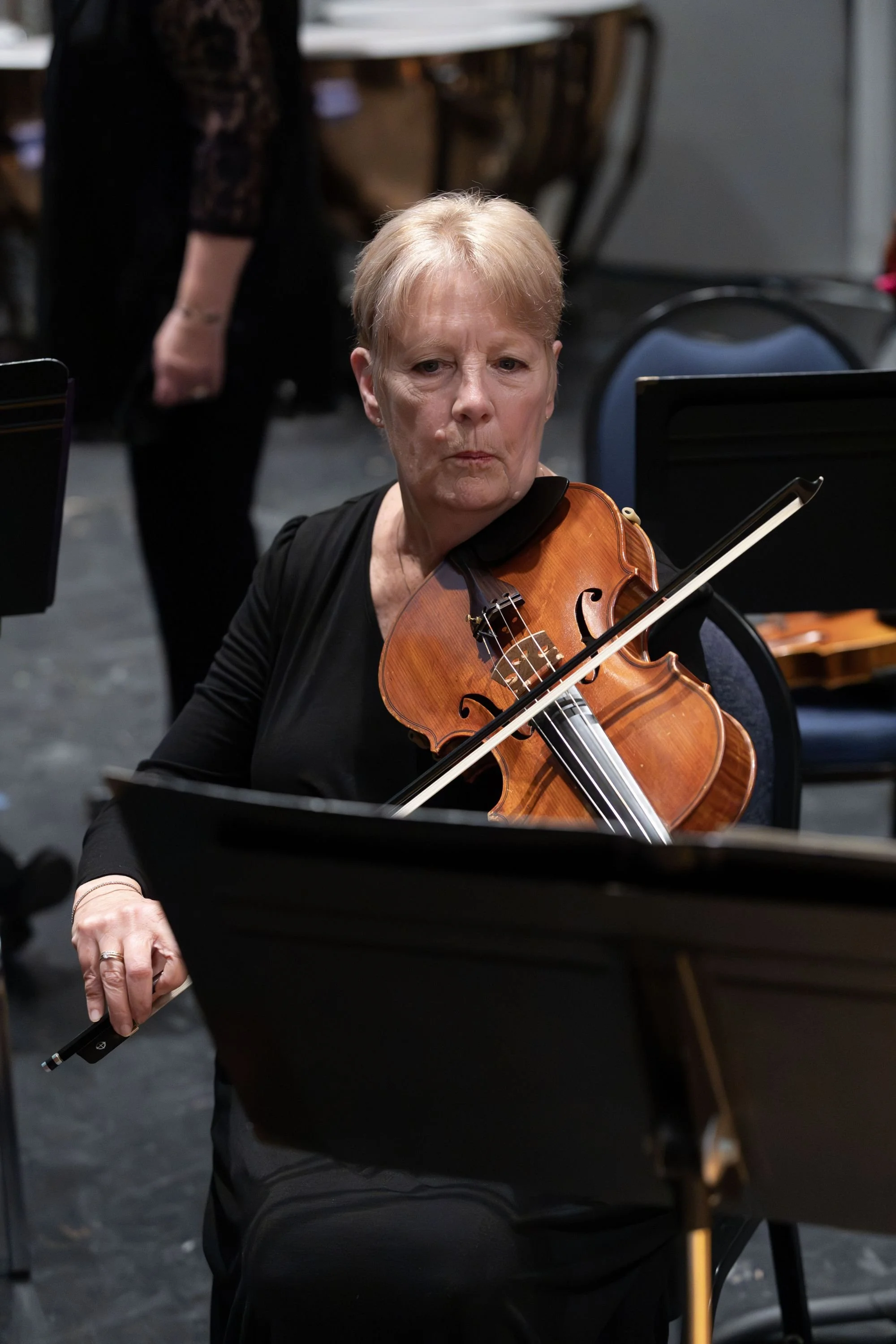 An elderly woman with short blonde hair, wearing a black top, is playing a violin during a rehearsal or concert, with sheet music stands and other musicians in the background.