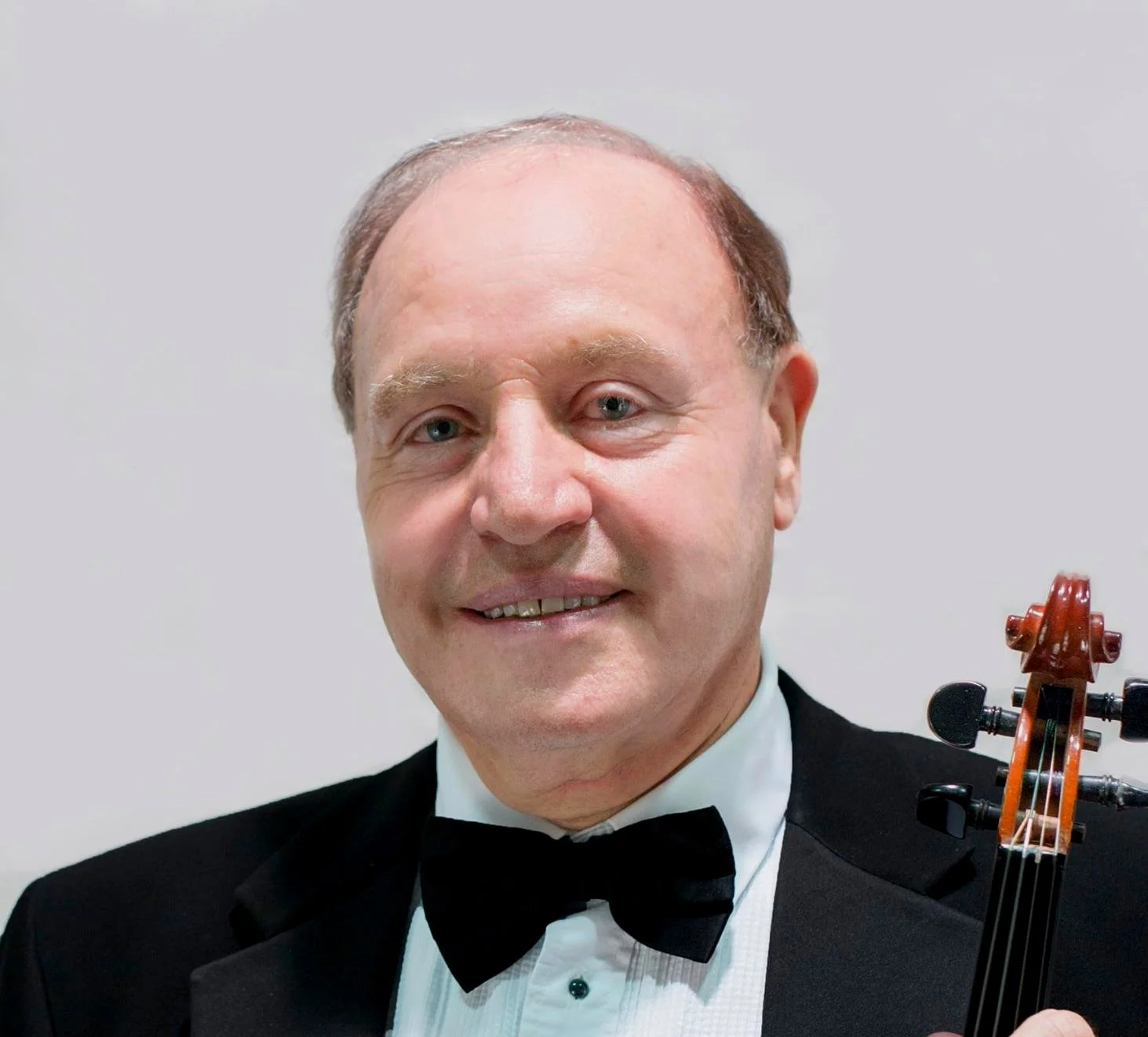 A man in a tuxedo and bow tie holding a cello. The man has light skin, short light brown hair, and blue eyes, standing against a plain white background.