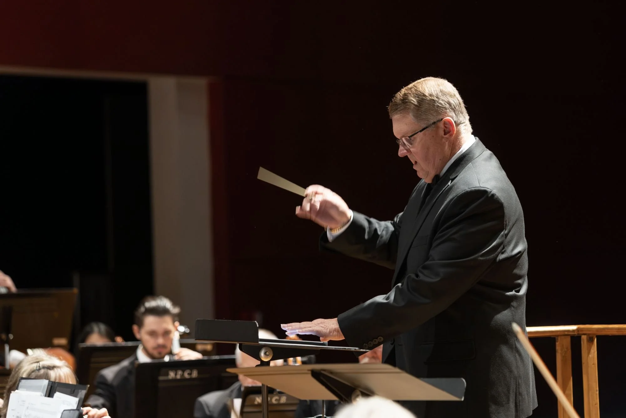 A male orchestra conductor in a black suit leading an orchestra, holding a baton in his right hand, with musicians and music stands visible in the background.