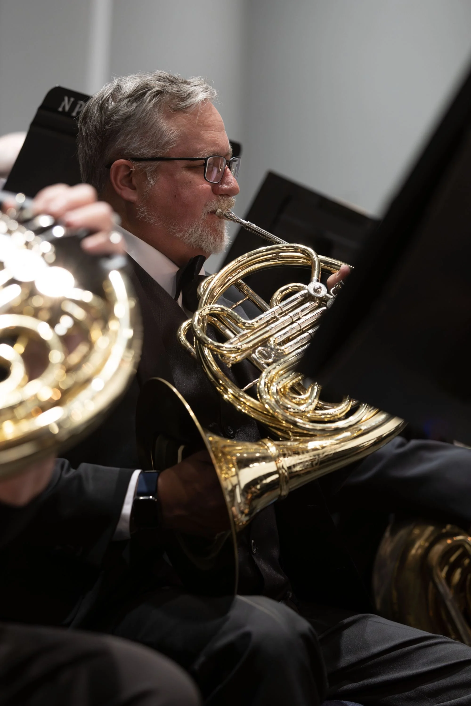 A man with glasses and a beard playing a French horn during a musical performance, with other musicians also playing brass instruments in the background.