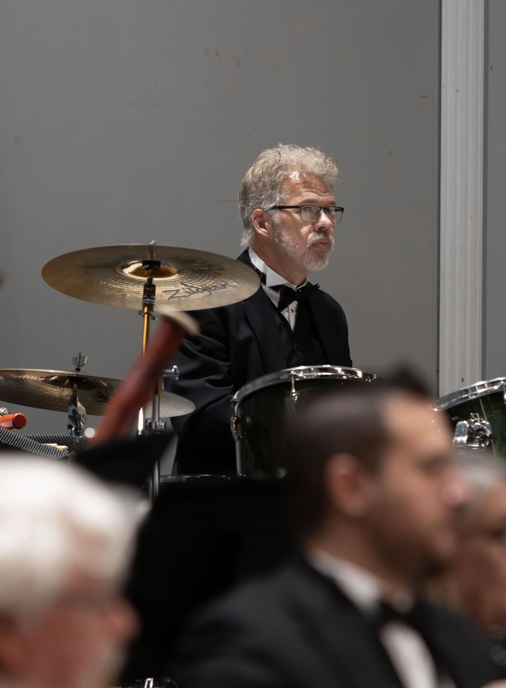 A middle-aged man with curly blond hair and glasses, dressed in a tuxedo, playing the drums at a formal event.