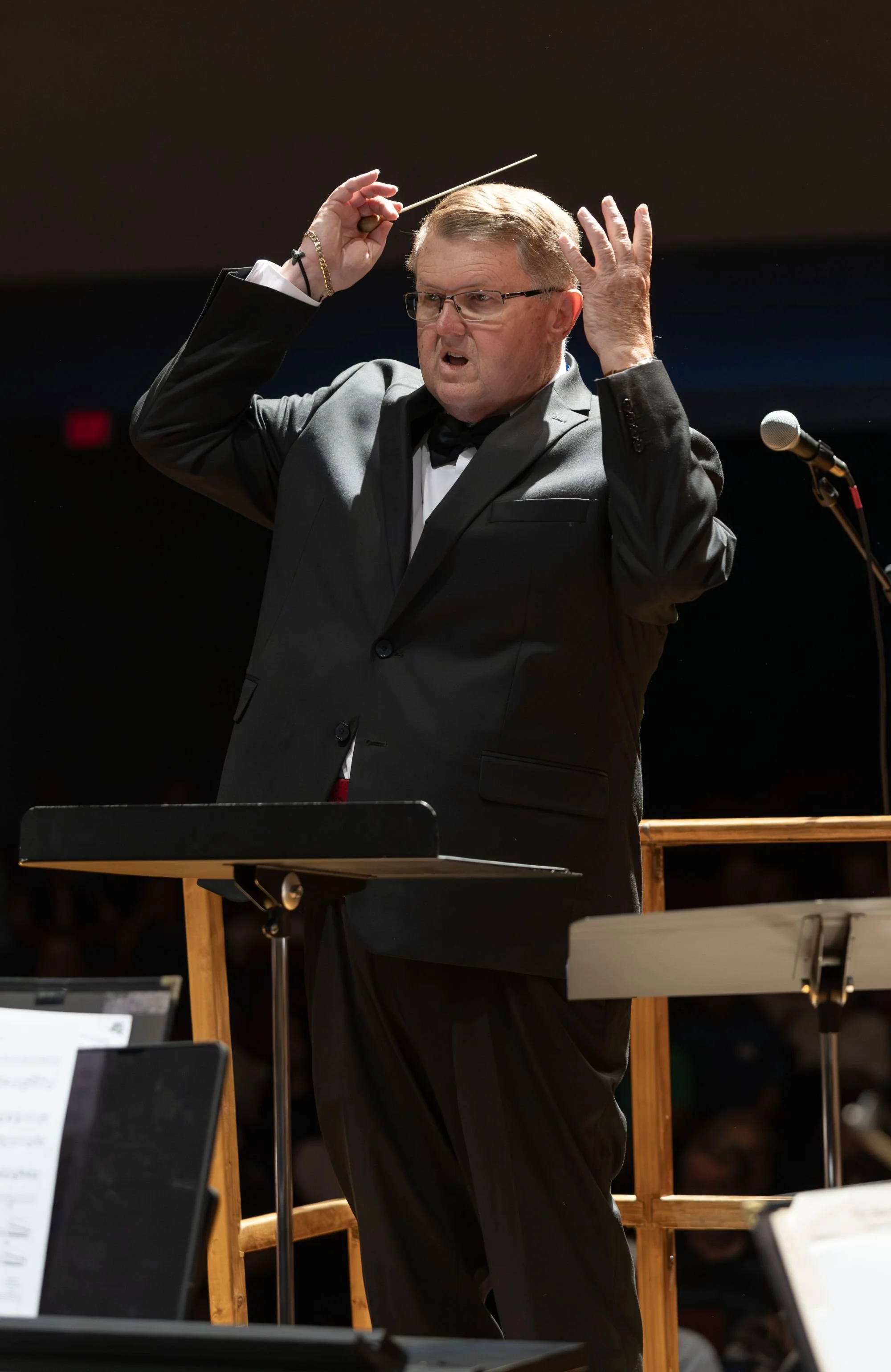 A man in a tuxedo conducts an orchestra, holding a baton above his head, with music stands and sheets in the foreground.