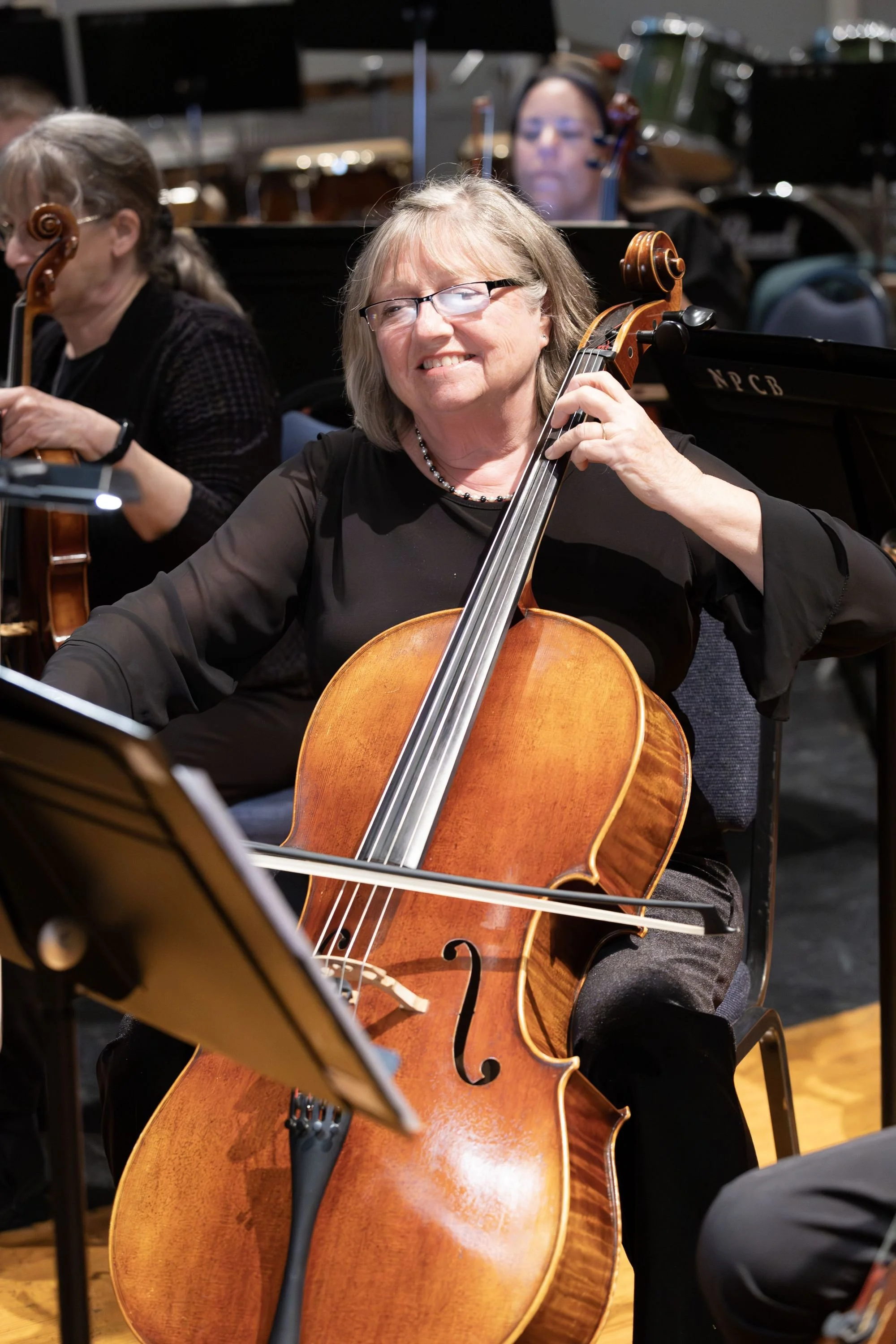 Musicians in an orchestra, with a woman playing the cello in the foreground and two other musicians in the background.