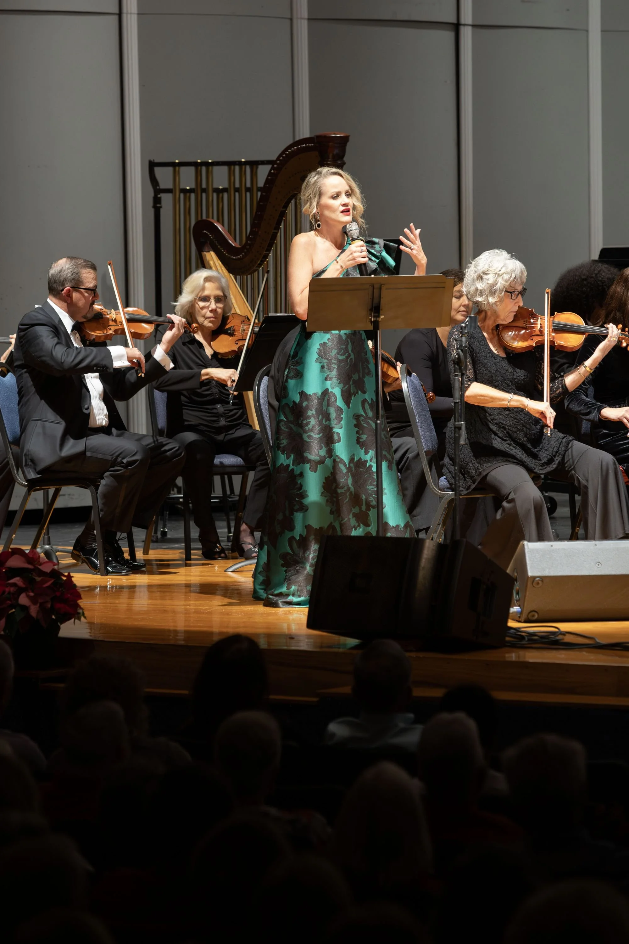 A woman in a green and black gown performing on stage with an orchestra, singing into a microphone while musicians play violins and a harp, with an audience watching.