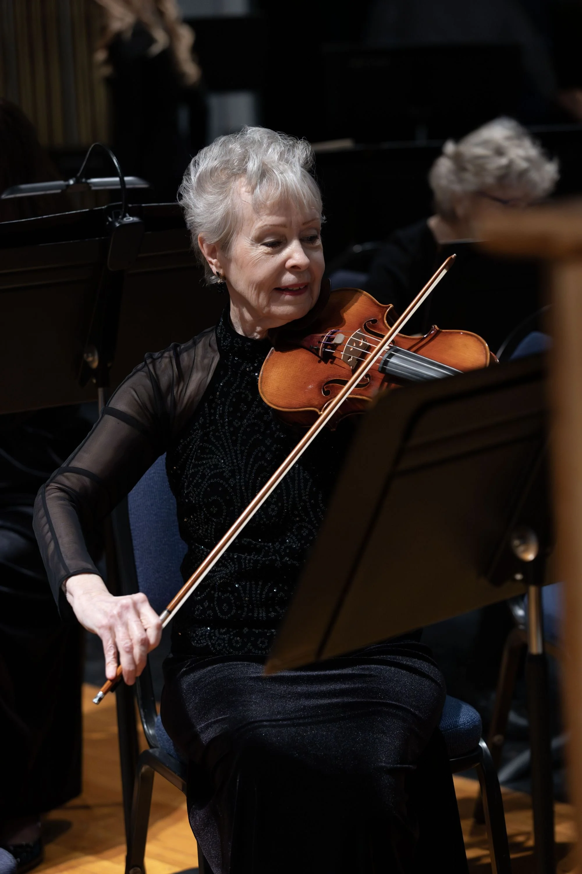 An elderly woman playing the violin during an orchestra rehearsal or performance.