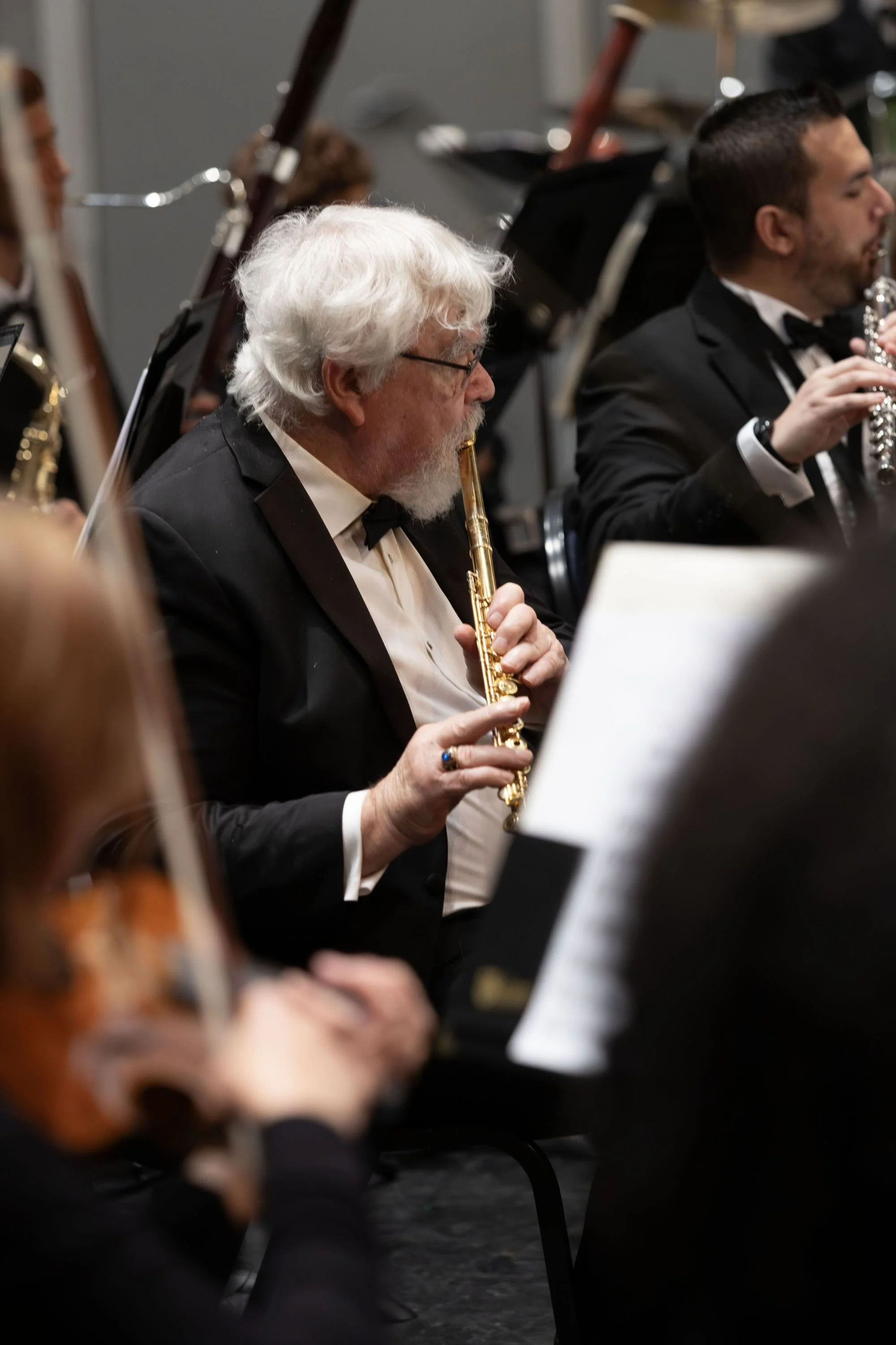 An older man with white hair and glasses playing a flute during an orchestra performance, with other musicians around him.