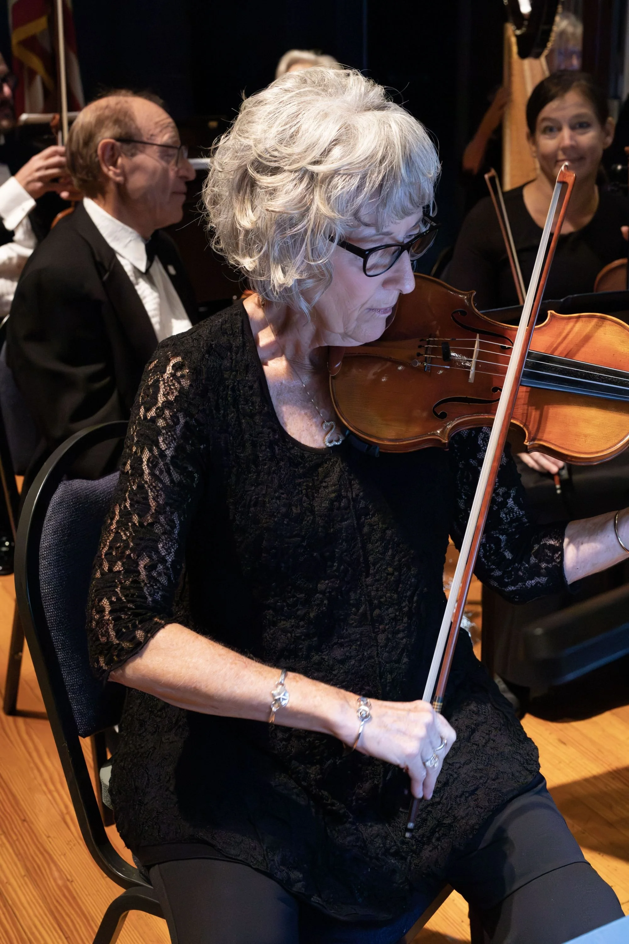 An elderly woman with gray hair and glasses playing the violin in an orchestra, surrounded by other musicians.
