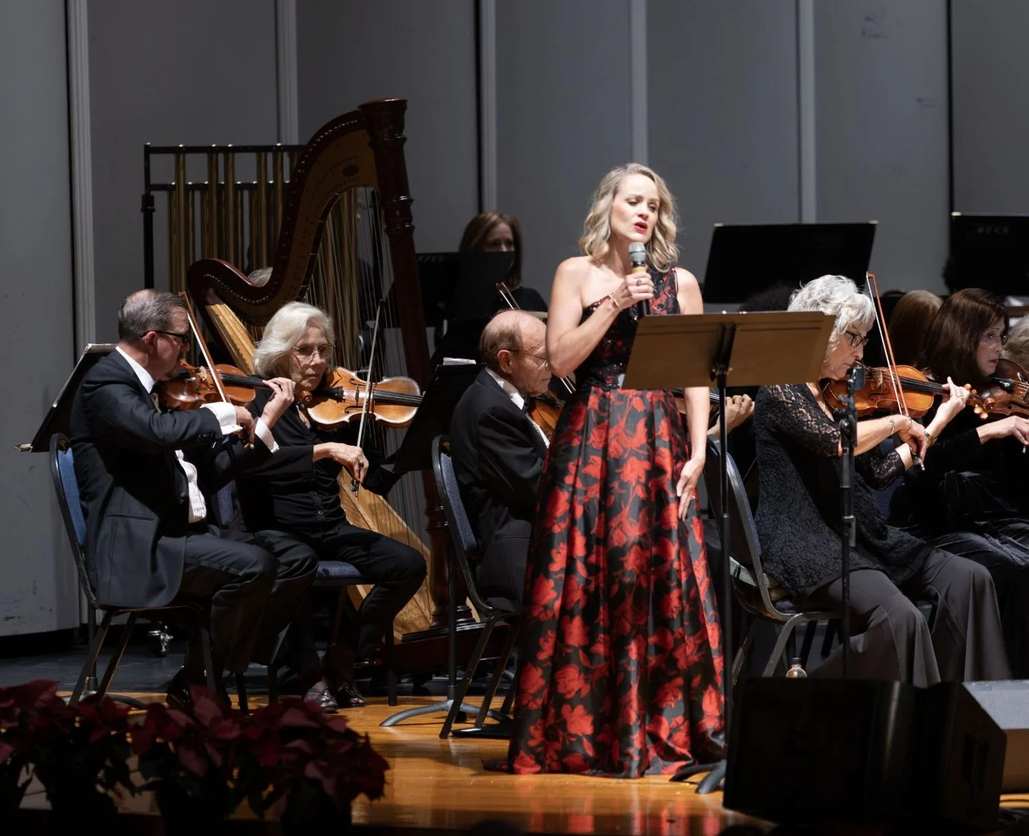 A woman in a black and red floral dress singing into a microphone on stage with an orchestra playing string instruments behind her.