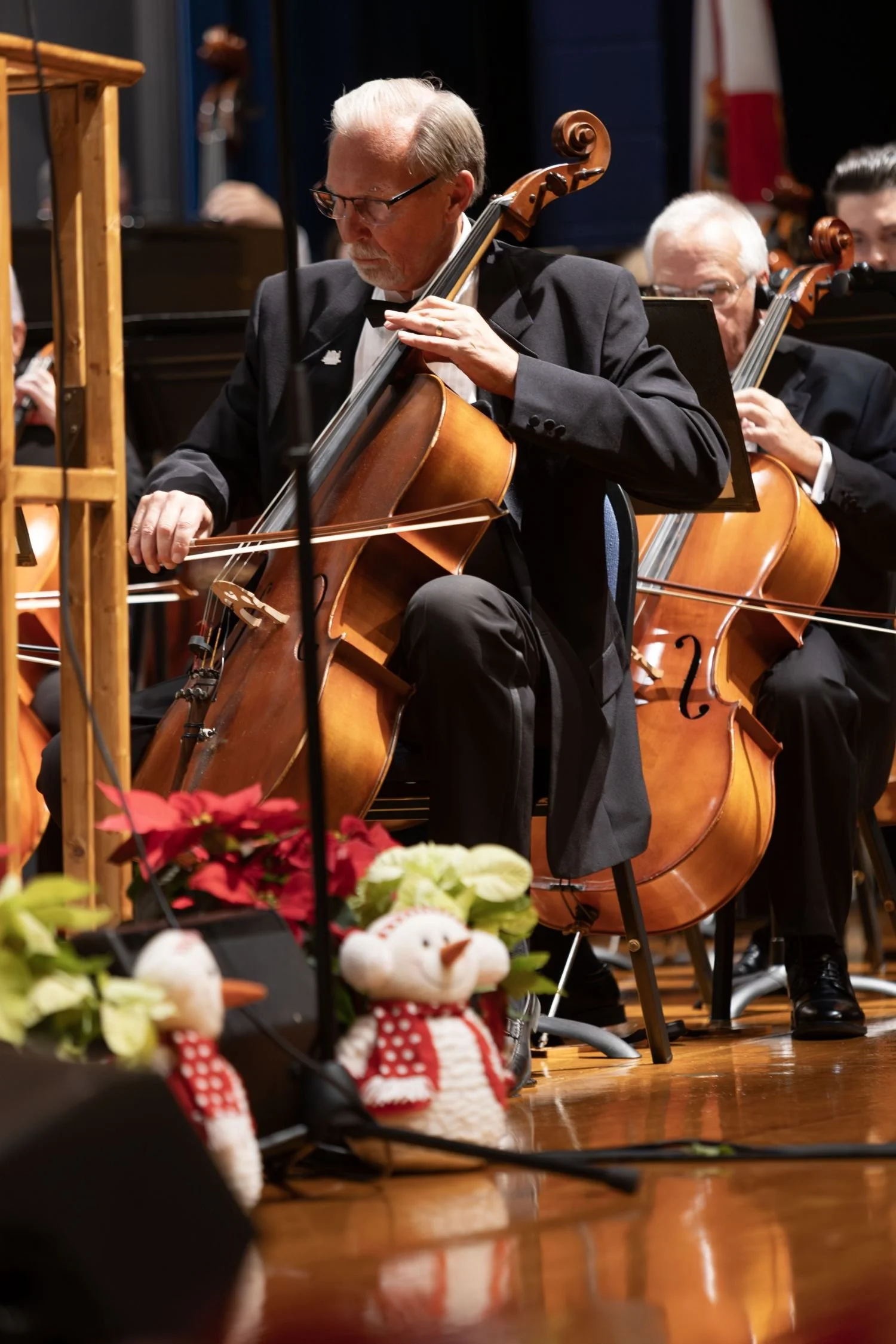 A group of musicians playing cellos during a Christmas concert, with festive decorations like poinsettias and snowmen plush toys in front of them.