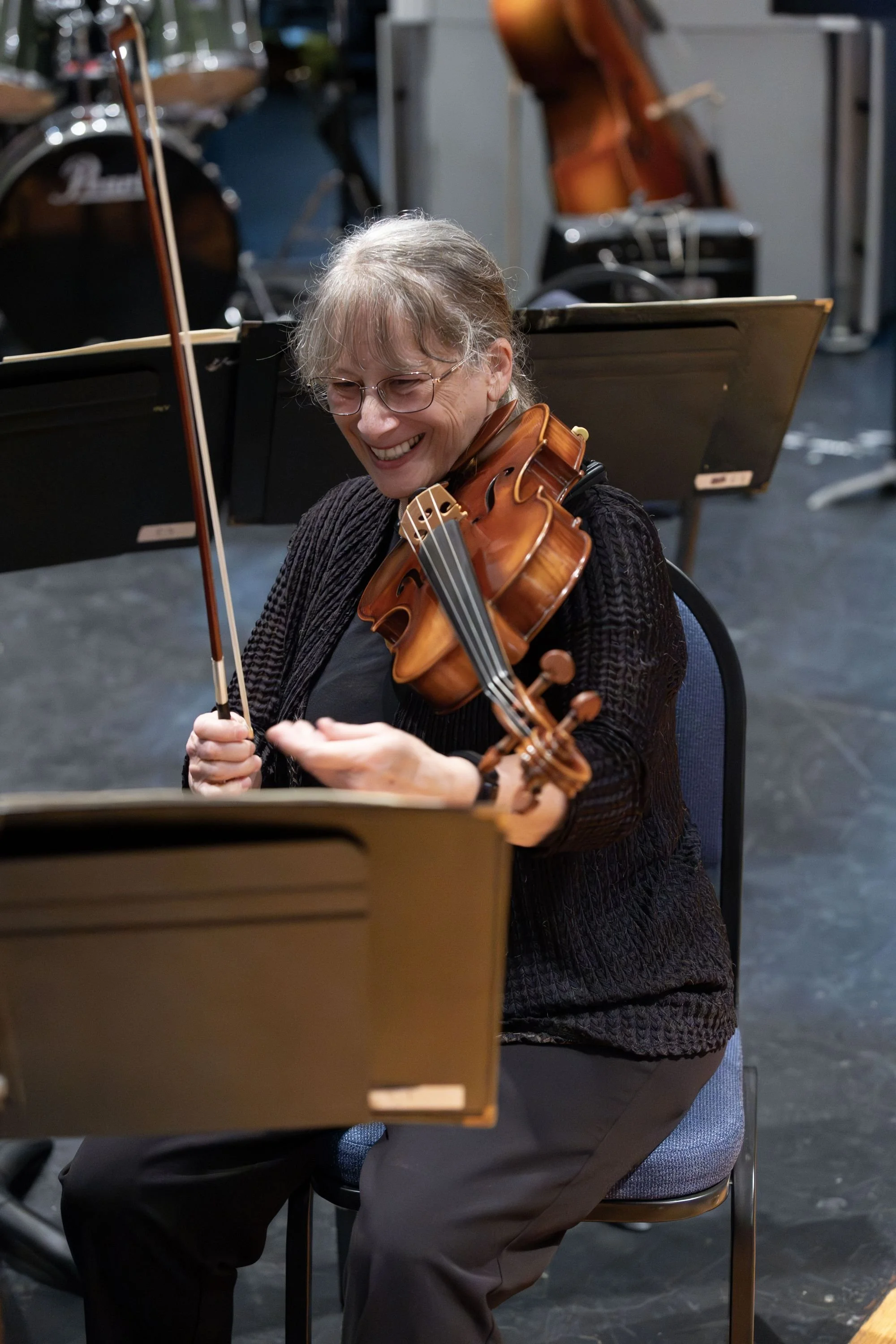 A woman with gray hair, glasses, and a black sweater playing a violin, smiling during a rehearsal with music stands and instruments around.