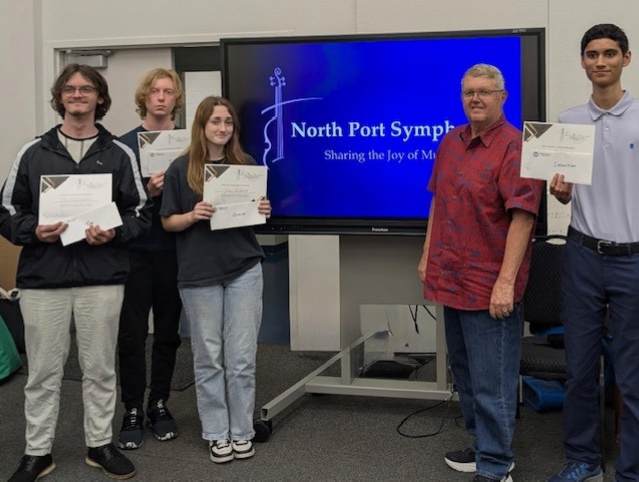 Group of five young students and one adult standing in front of a screen that displays "North Port Symphony" at an awards or certificate ceremony, each holding certificates.